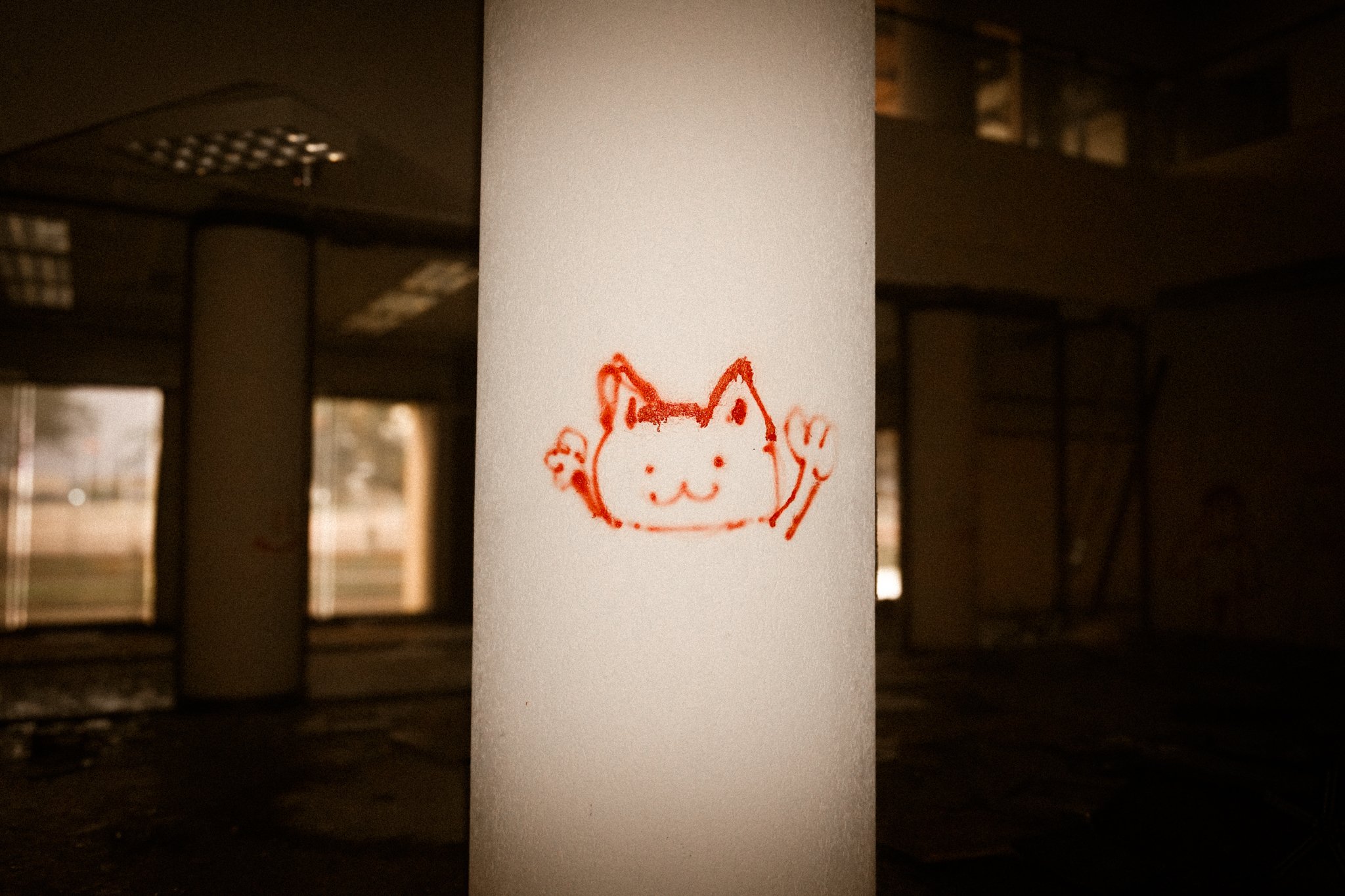 A red marker drawing of a cartoon cat with raised paws on a white pillar in an empty, dimly lit, abandoned building with large windows in the background.