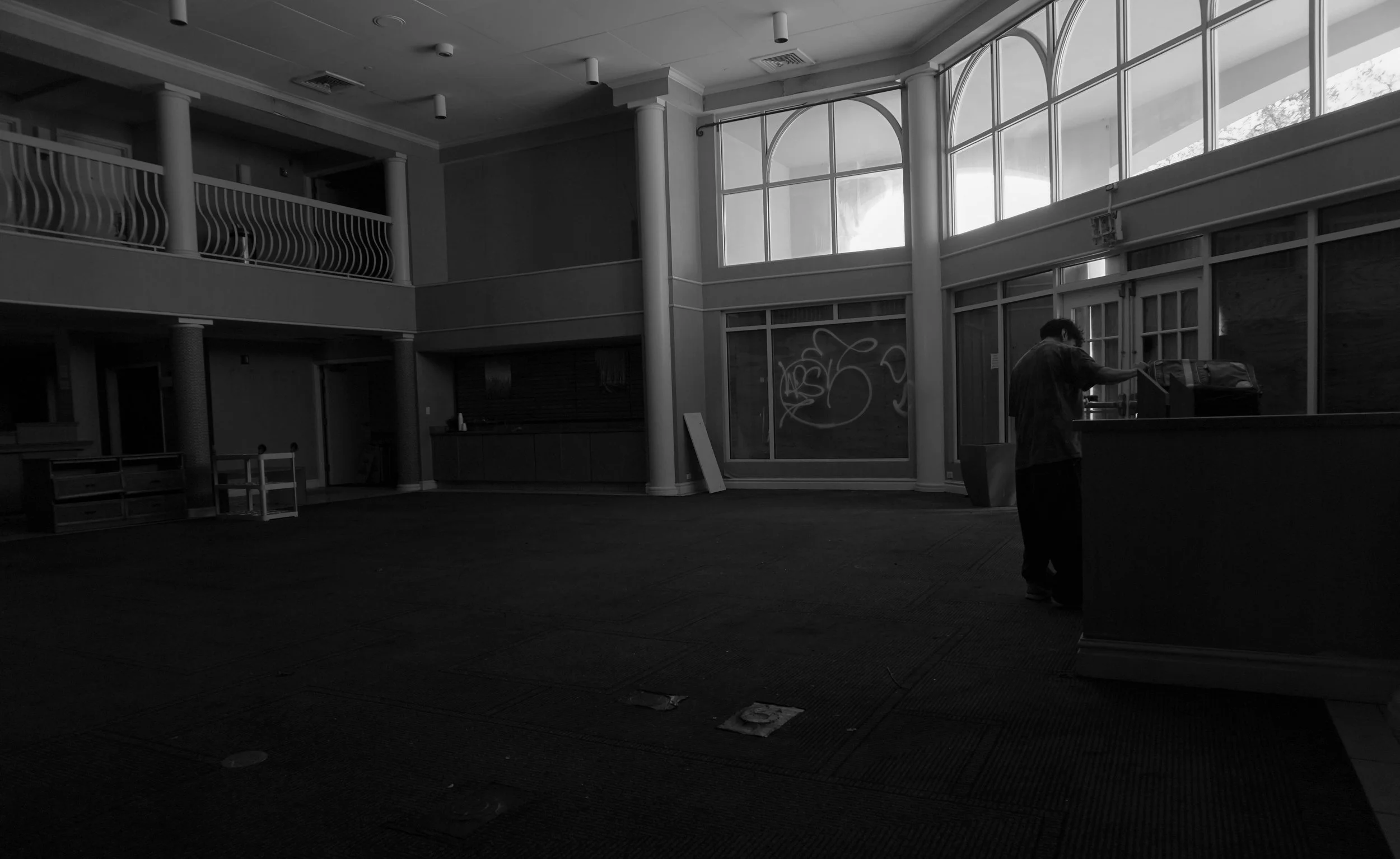 An empty indoor space with large windows, a person leaning over a counter, and graffiti on the glass wall.