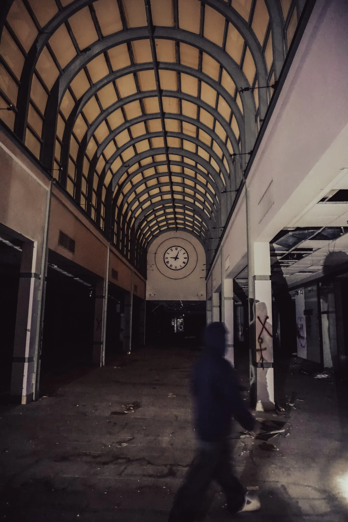 An abandoned shopping mall with a large arched glass ceiling and a clock on the back wall showing approximately 1:15. The corridor is dark and empty, with debris on the floor and graffiti on some walls. A person is walking through the space.