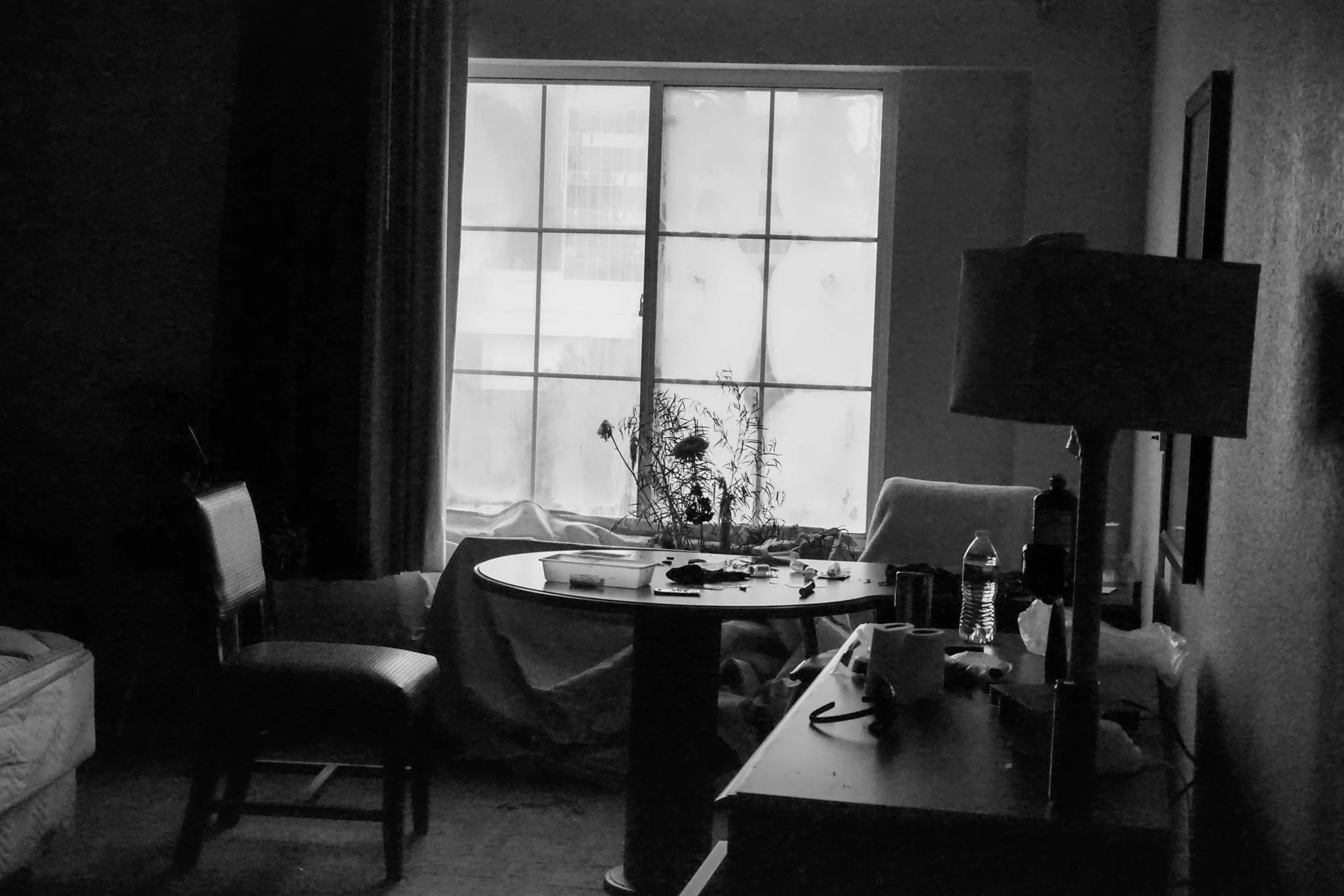 Dimly lit hotel room with a round table, chairs, a window, and scattered objects like a water bottle, papers, and a lamp.