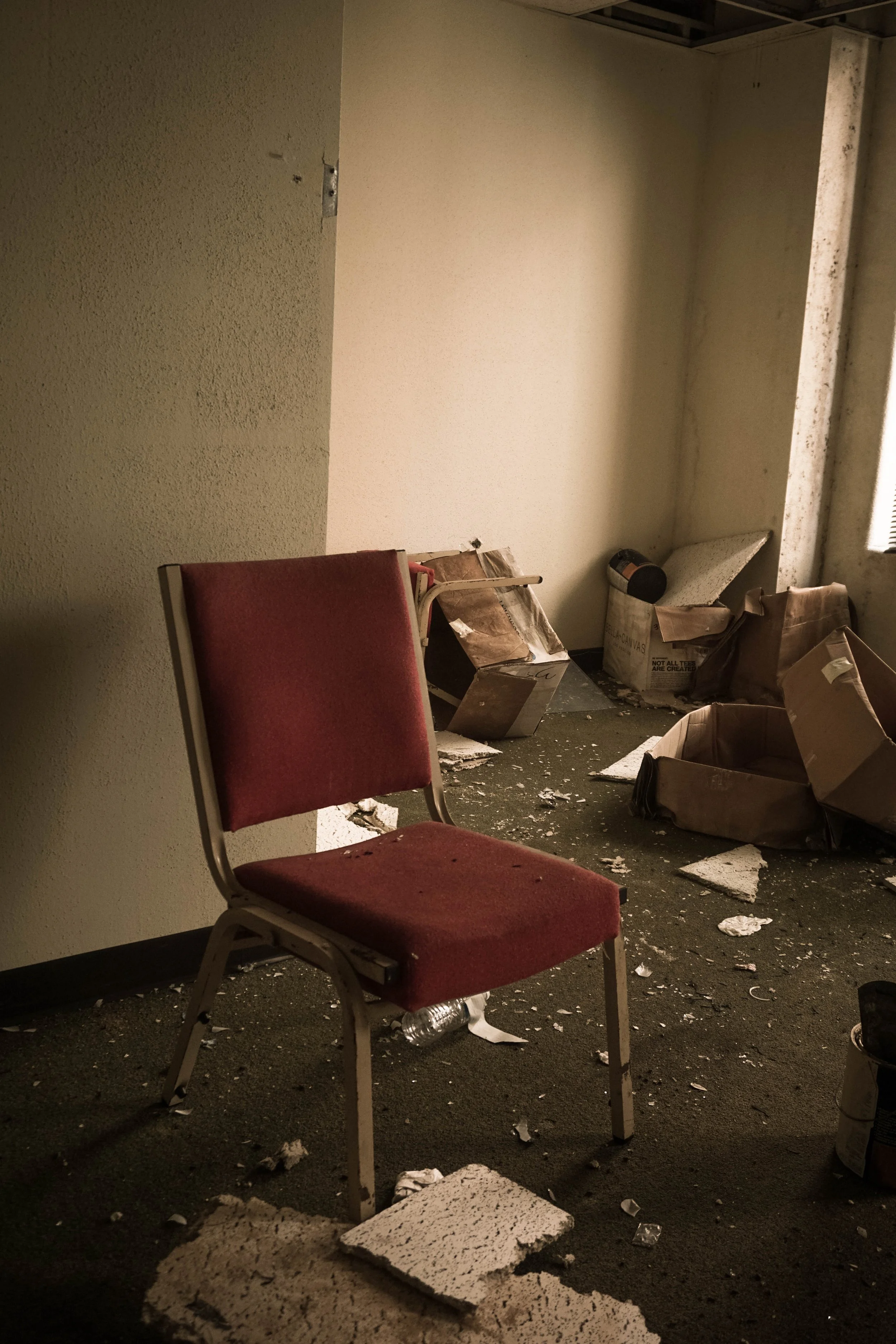 An abandoned room with a red padded chair and debris, including broken drywall and cardboard boxes, scattered on the floor.