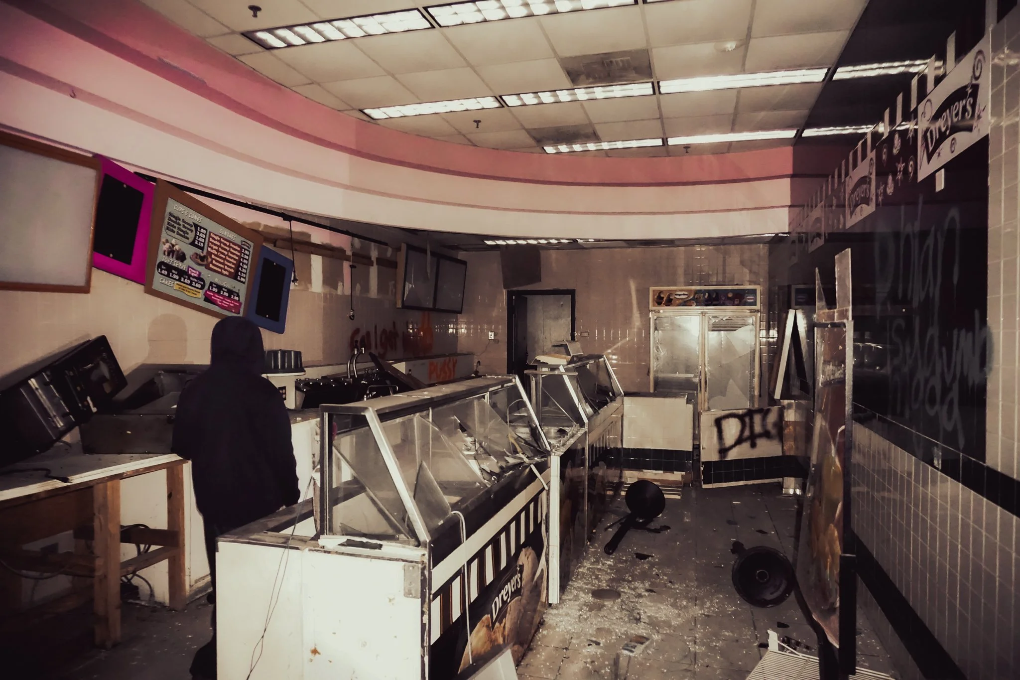 Interior of a fast food restaurant with damaged counters, broken speakers on the floor, and graffiti on the walls, with a person wearing a black hoodie standing among the debris.