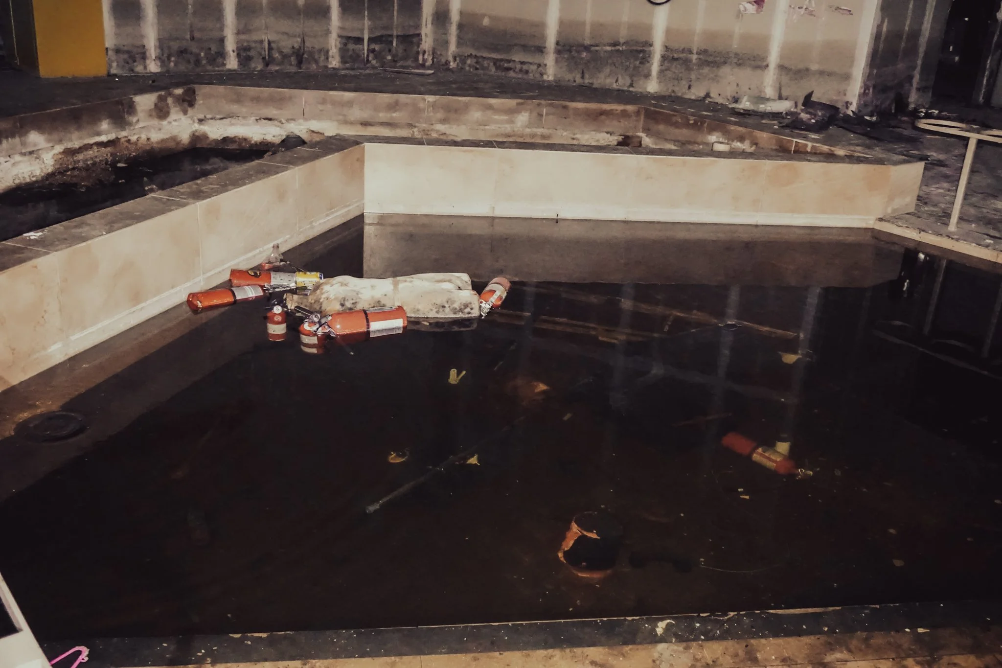 An indoor swimming pool under construction or renovation with debris, water, and fire extinguishers floating in the water. The area appears damaged, with missing fixtures and construction materials scattered around.