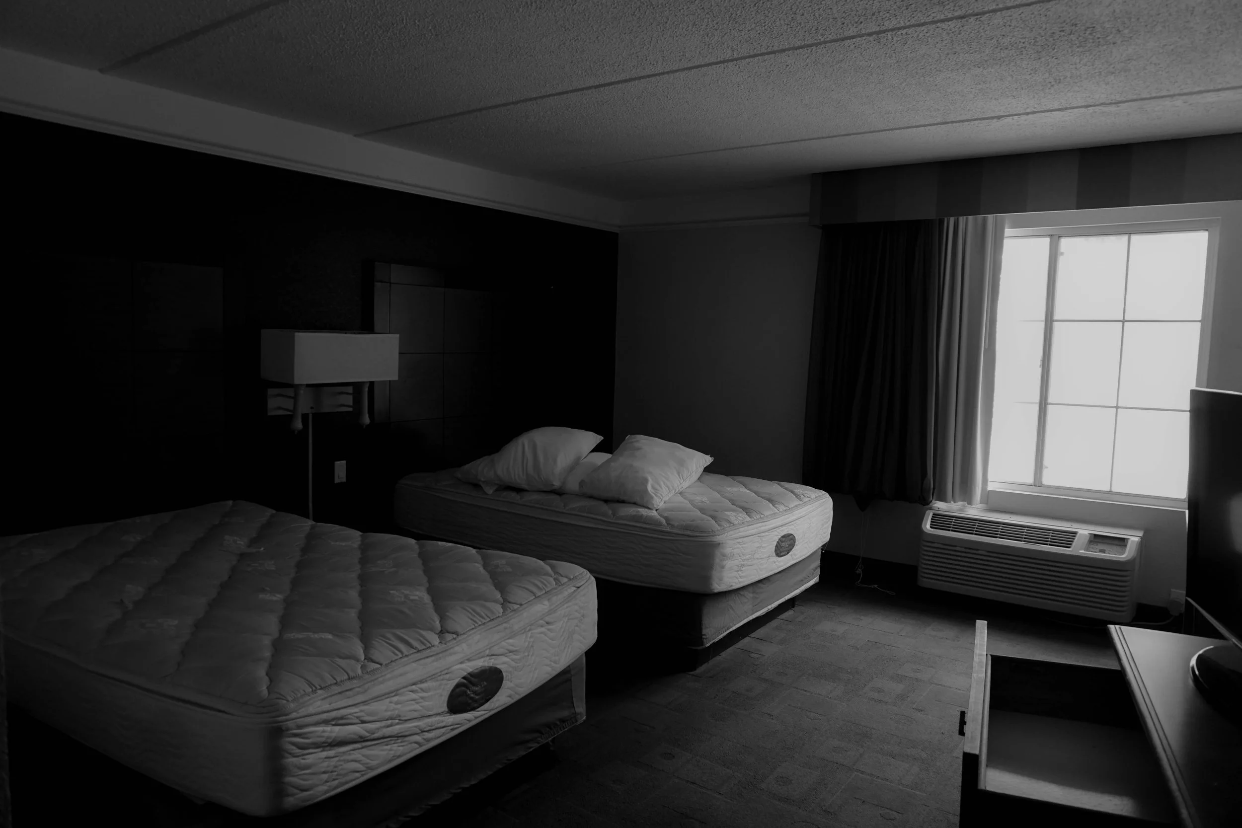 A hotel room with two beds, one bed with three pillows, a window with closed curtains, an air conditioning unit under the window, and a small table or dresser in the foreground.