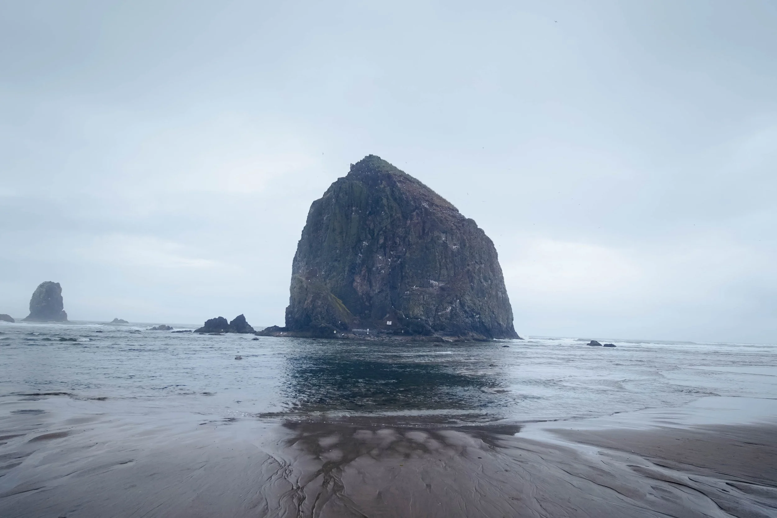 A large sea stack rock formation emerging from the ocean near a sandy beach, with smaller rocks nearby under a cloudy sky.