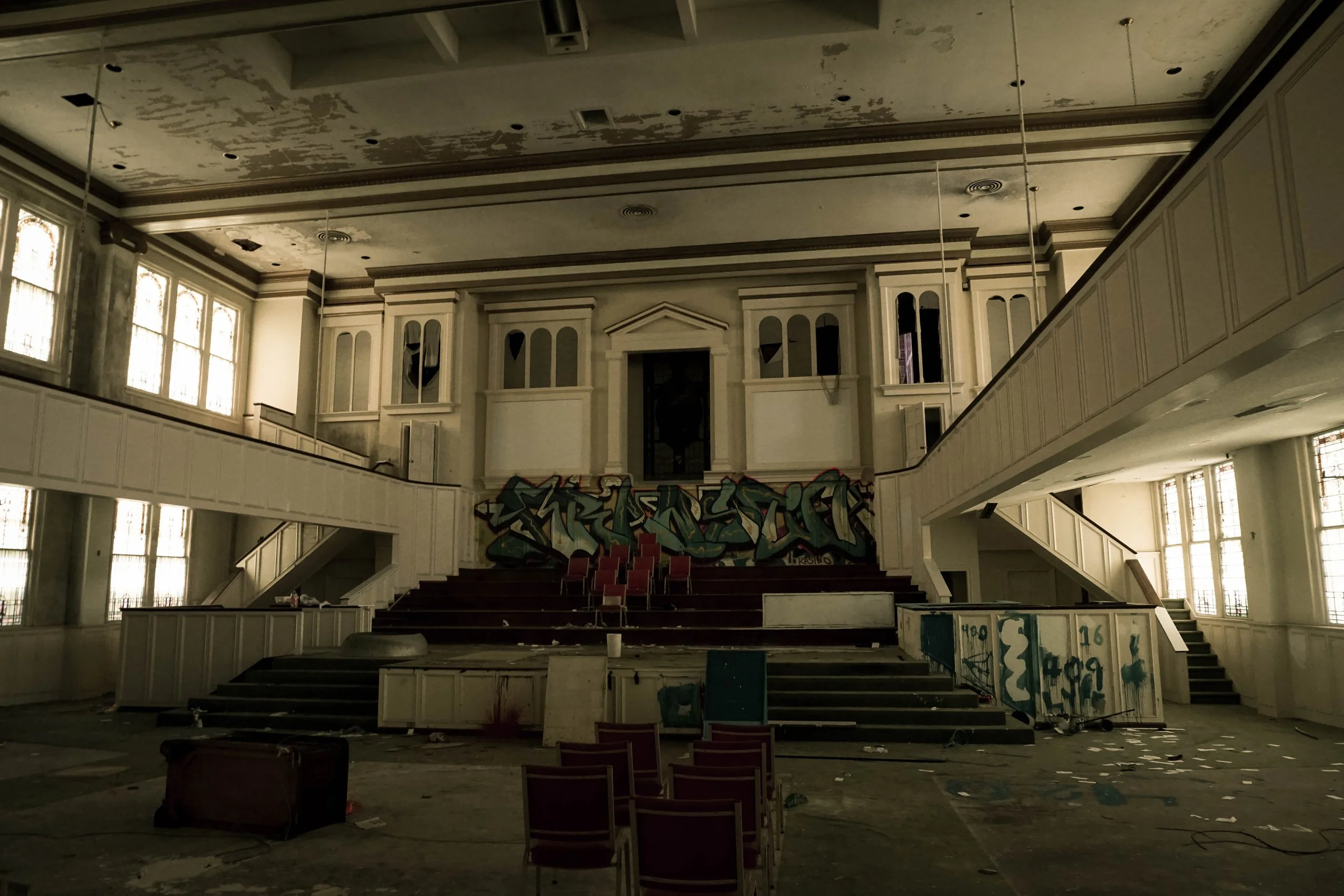 Interior of an abandoned building with a stage, graffiti, broken chairs, peeling paint, and debris scattered on the floor.