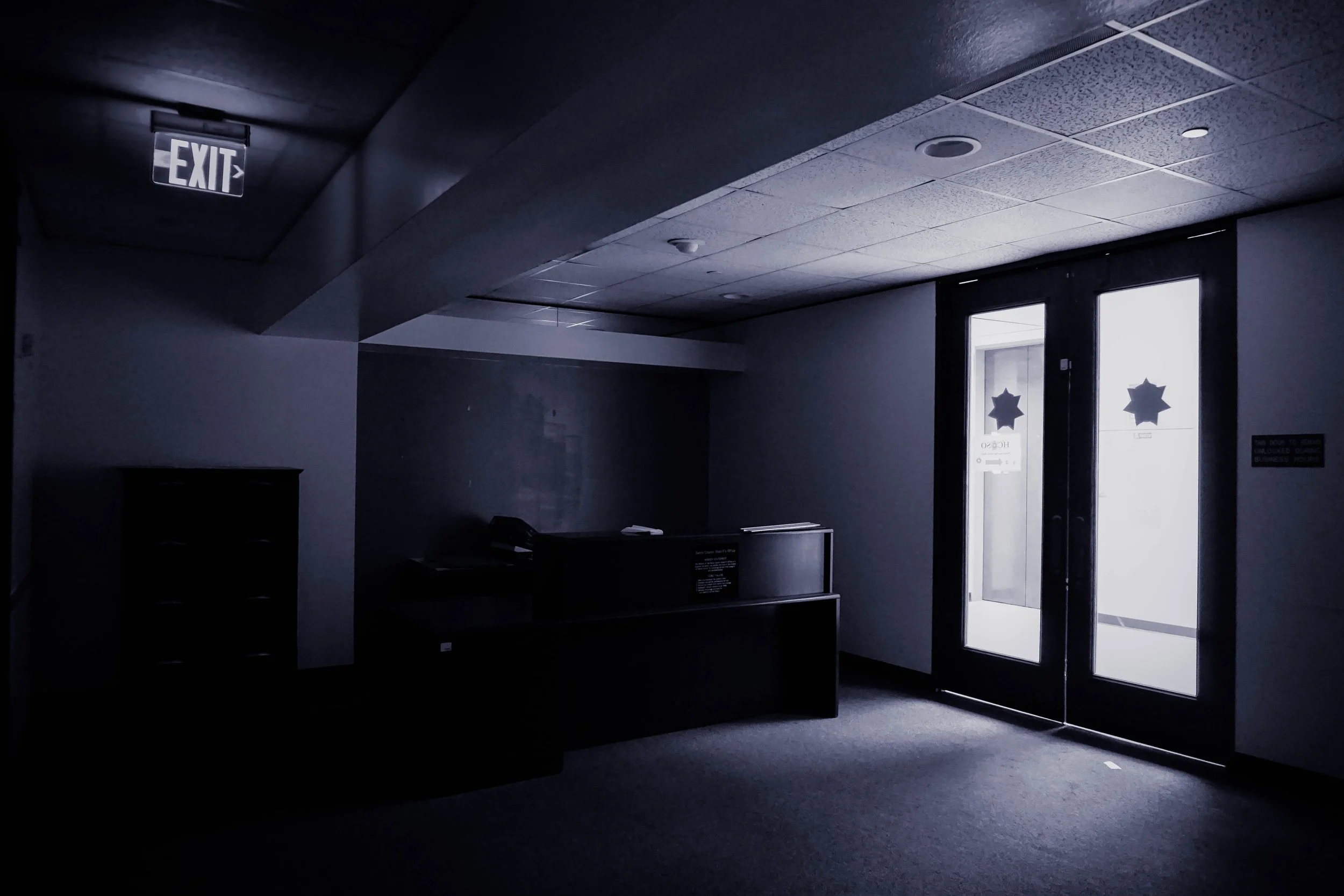 Dimly lit interior space with a front desk, a closed cabinet, and double glass doors with star-shaped security stickers, illuminated by bright light from outside.