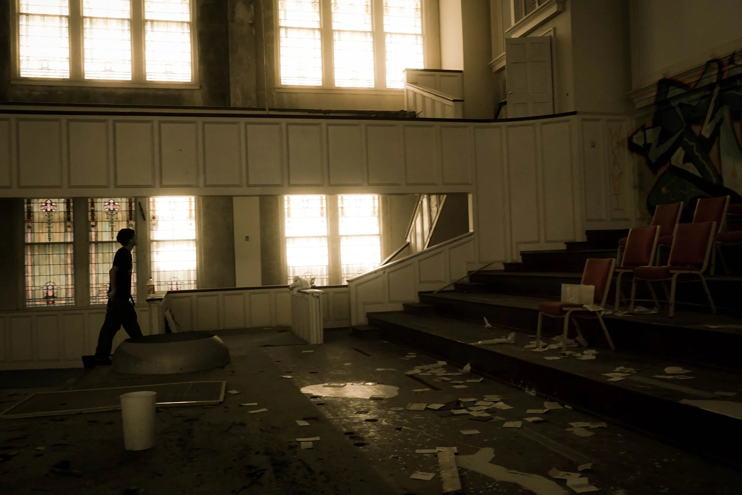 An abandoned building interior with scattered papers and debris on the floor, several empty chairs on the right side, a person walking along the left side near the window, and sunlight shining through large, stained-glass windows.