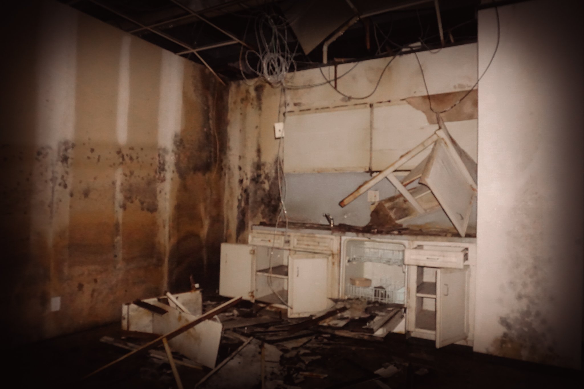 Deteriorated room with moldy walls, debris, and broken furniture including a damaged cabinet and overturned chair.