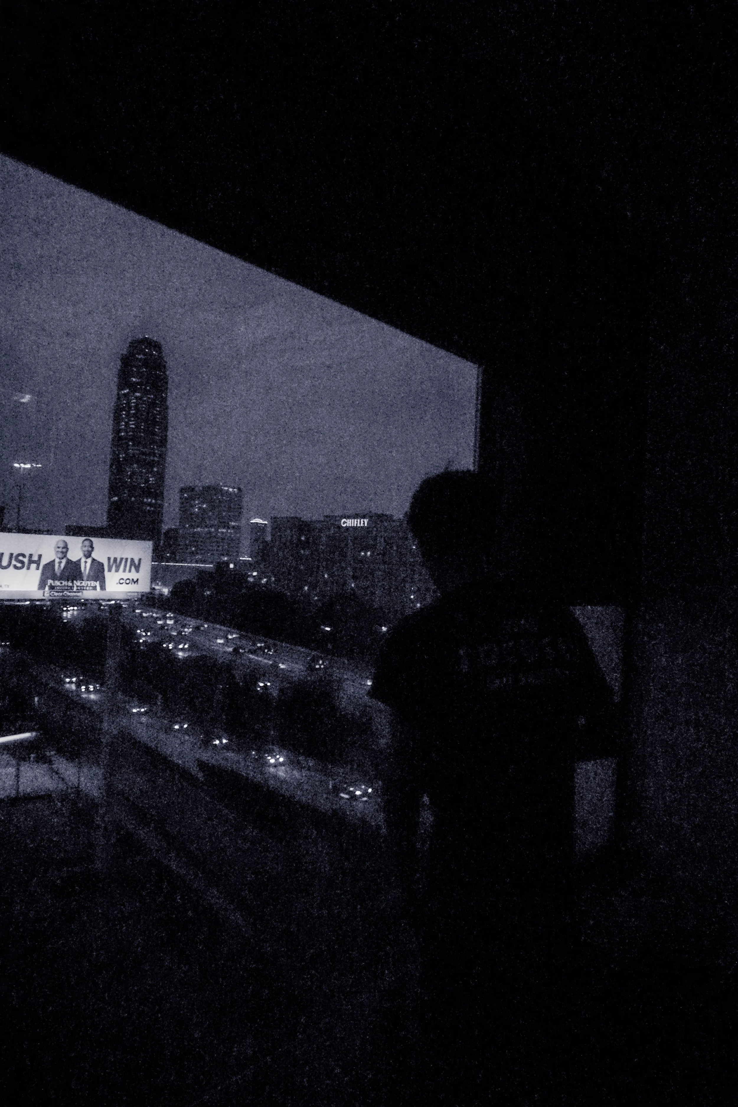 A person standing on a balcony at night overlooking a city skyline with illuminated buildings, traffic, and a billboard.