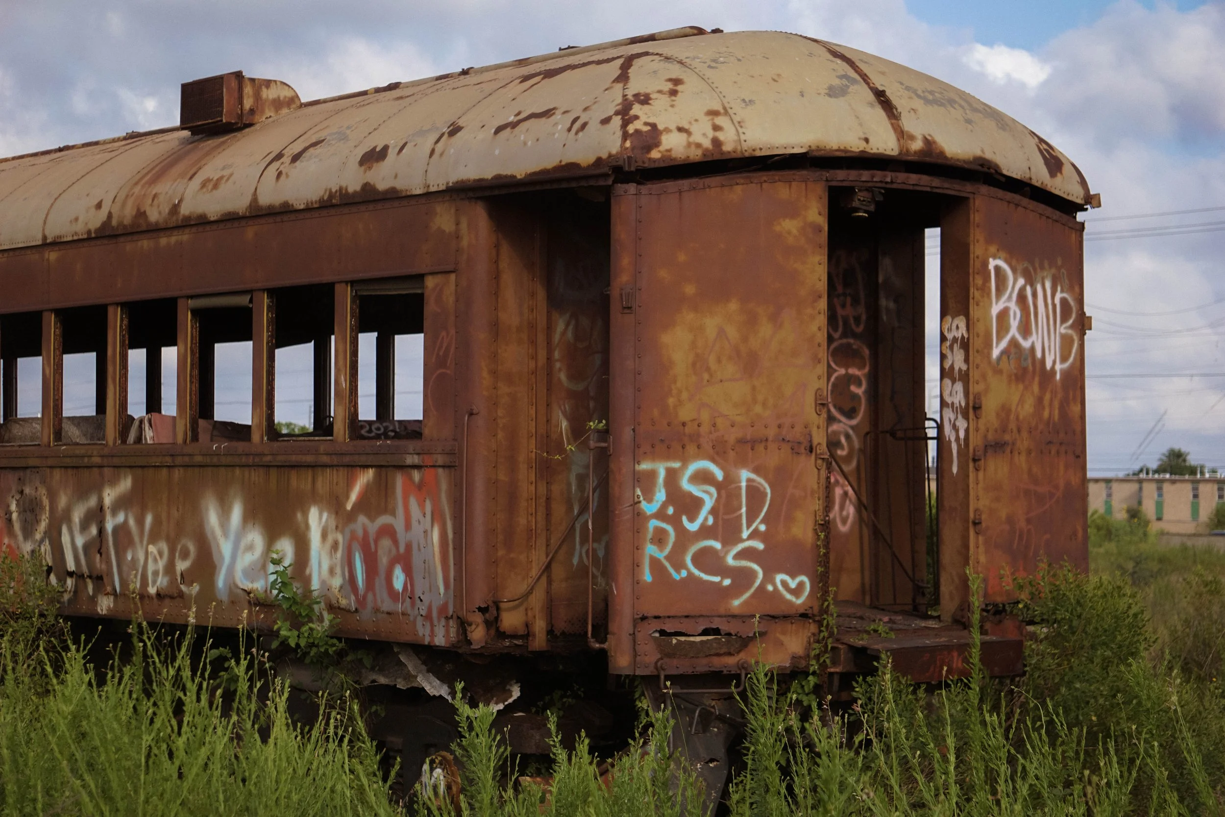 An old, rusted train car with graffiti, overgrown grass and plants surrounding it, under a partly cloudy sky.