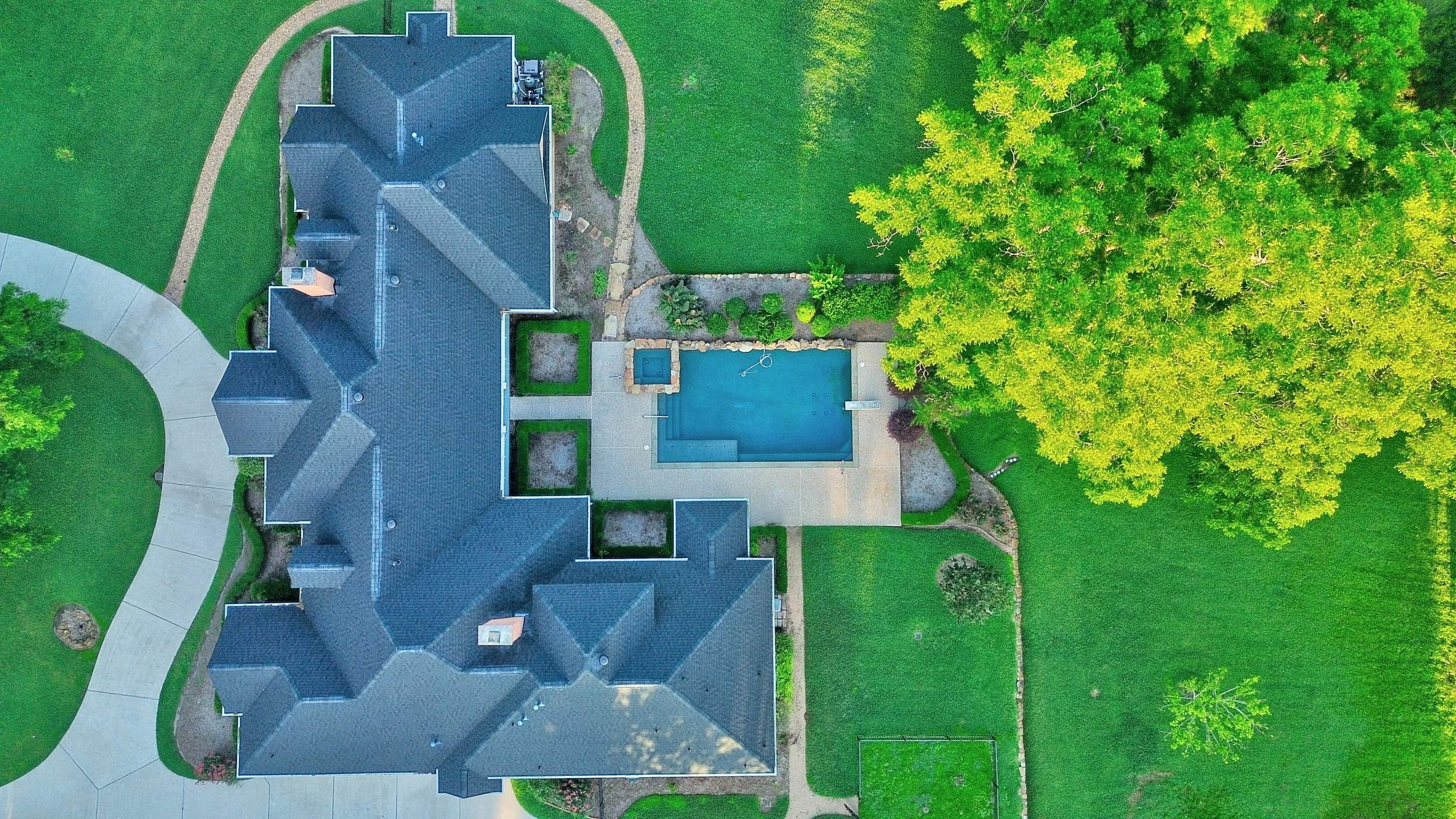 Aerial view of a house with a backyard featuring a swimming pool, surrounded by green grass, trees, and pathways.