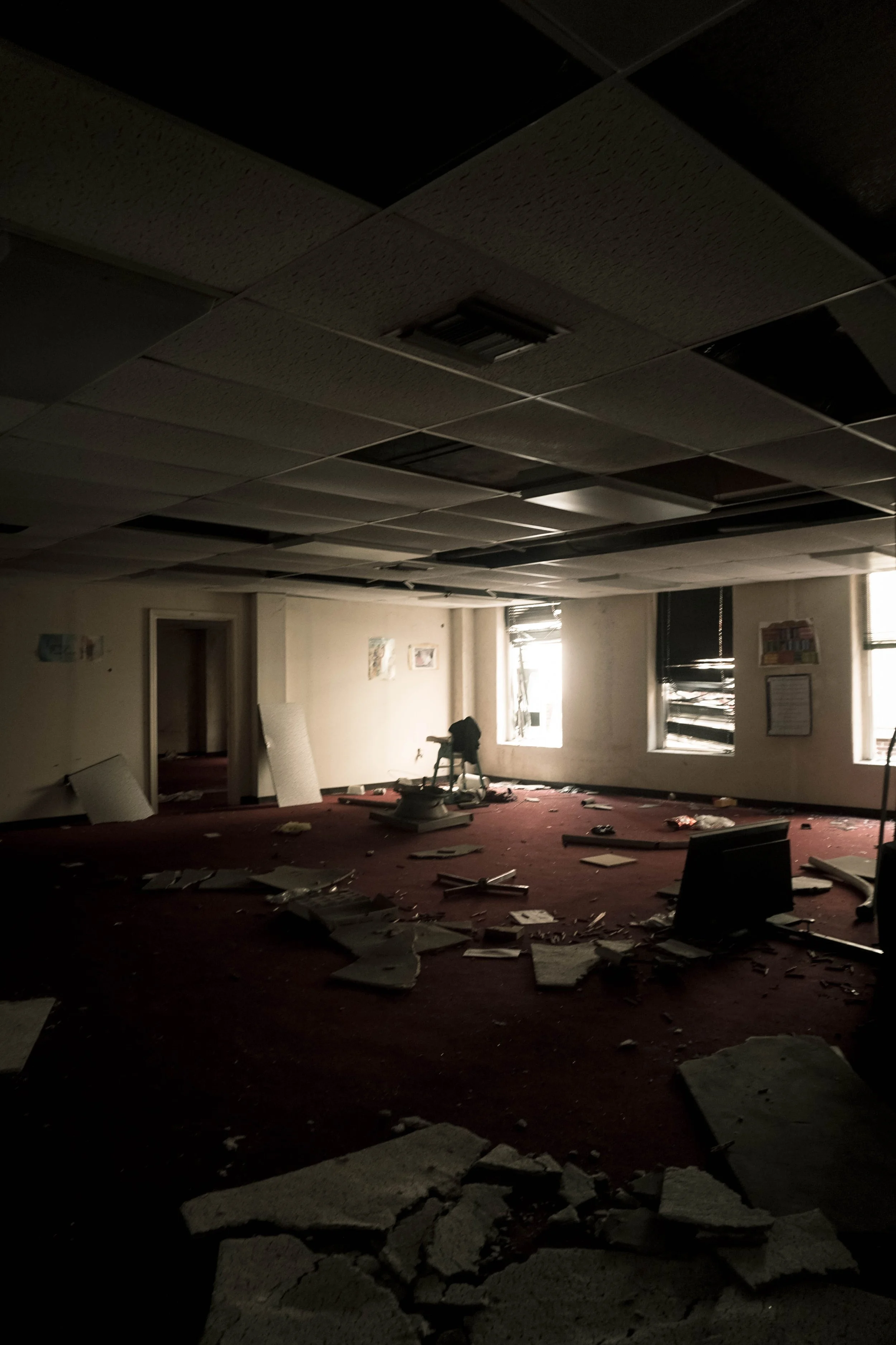 An abandoned room with debris scattered on the floor, damaged ceiling tiles, and broken windows letting in light.
