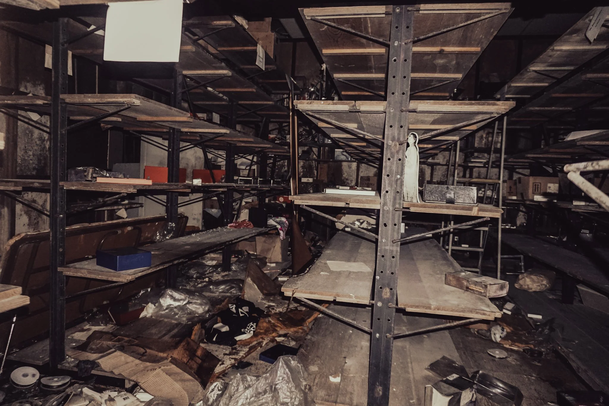 Dark, cluttered storage room with empty shelves, scattered debris, and a small religious figurine.