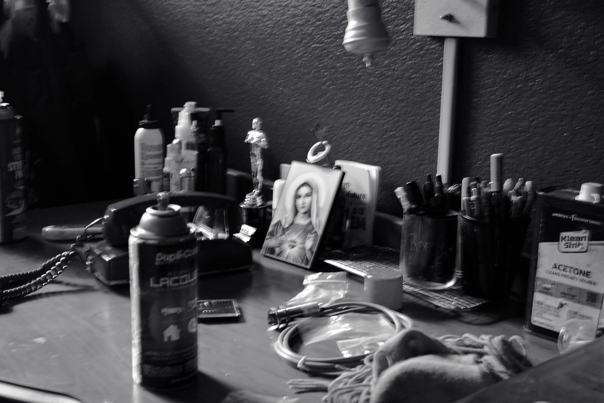 A cluttered desk with various bottles, pens, a framed picture of a woman, a dispenser box of acetone, a coiled cord, and miscellaneous items in black and white.