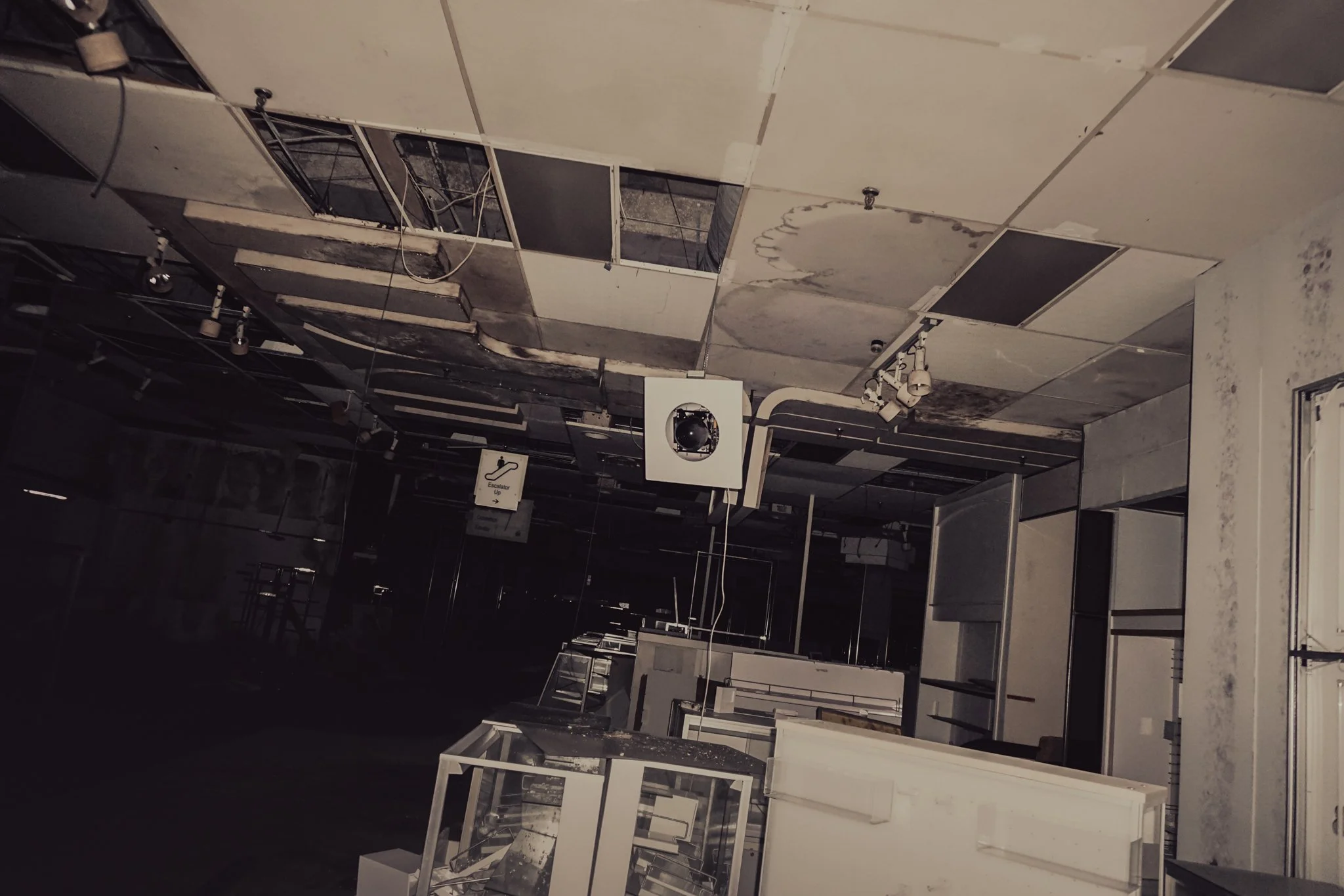 An abandoned and decaying retail store with damaged ceiling tiles, missing light fixtures, and broken display cases.