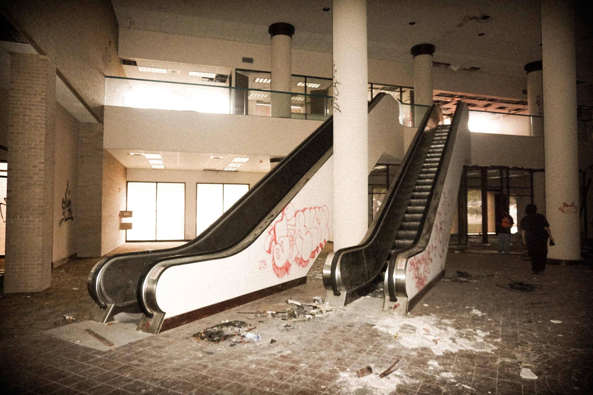 An abandoned indoor shopping mall or commercial space with two broken escalators, graffiti on the walls, scattered trash on the floor, and two people walking in the background.