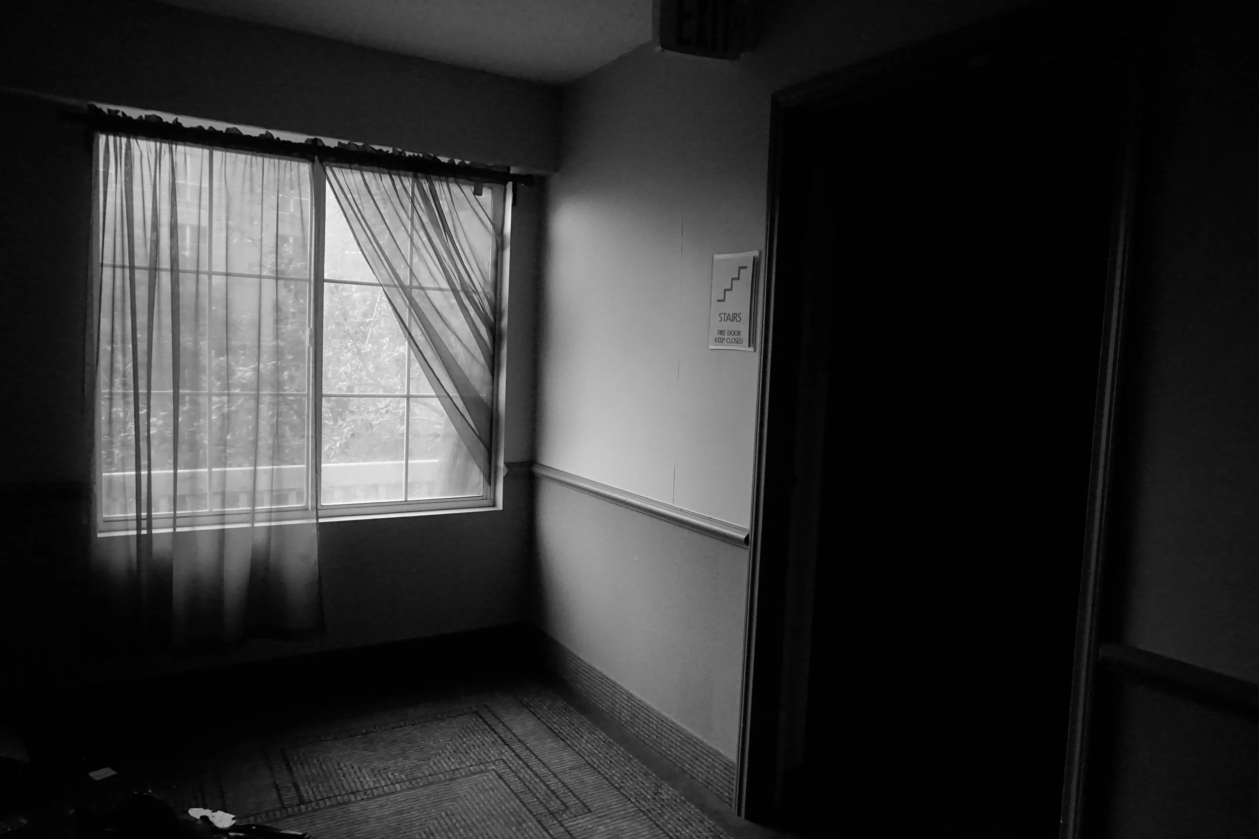 A black and white photo of an indoor space with a window covered by flowing curtains, a wall with a staircase sign, and an open doorway leading to darkness.