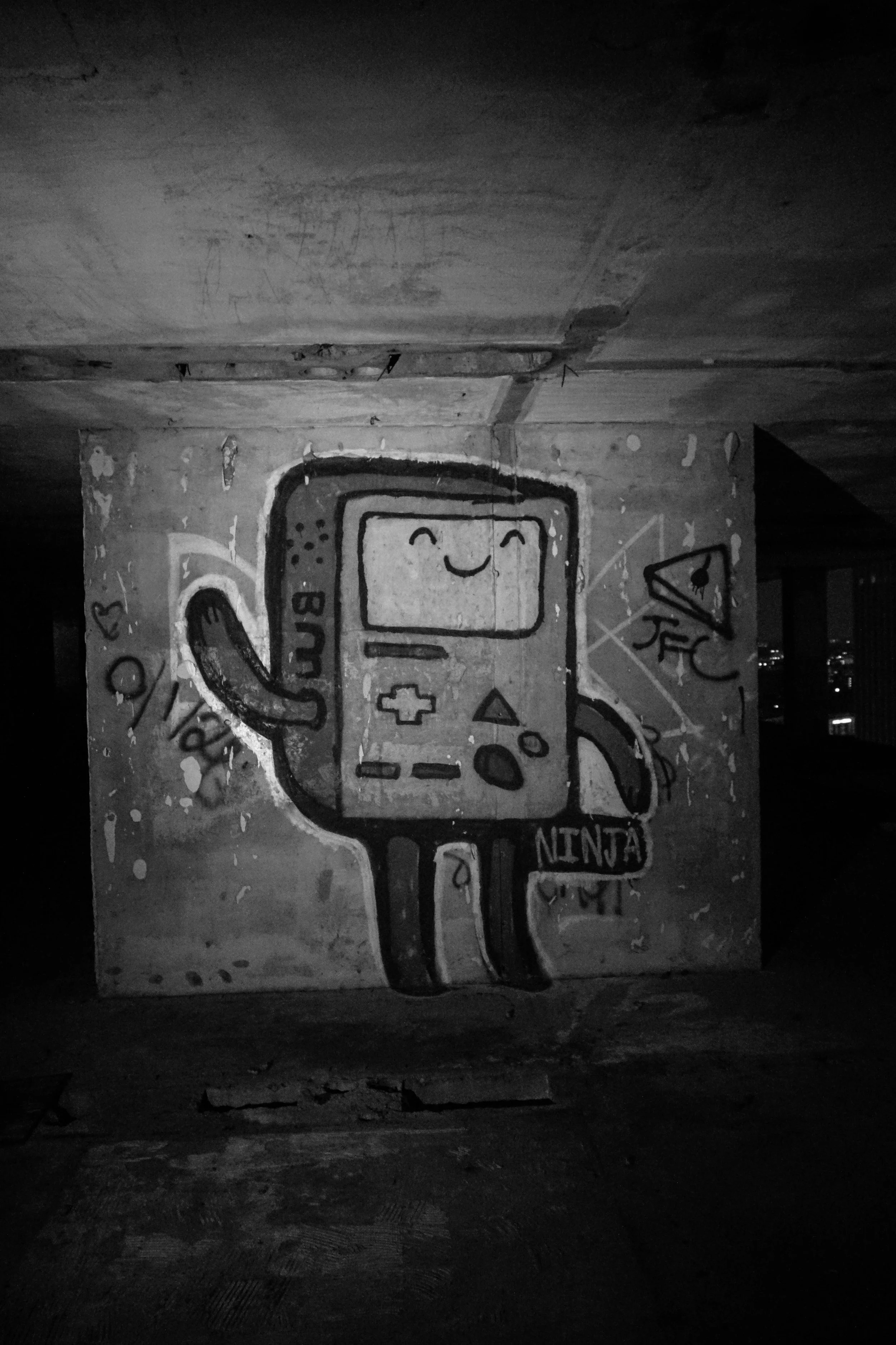 Graffiti of a cartoon character with a smiling face, resembling a handheld gaming console, with arms and legs, holding a bag labeled "NINJA." The graffiti is on a rough wall under a dark ceiling.