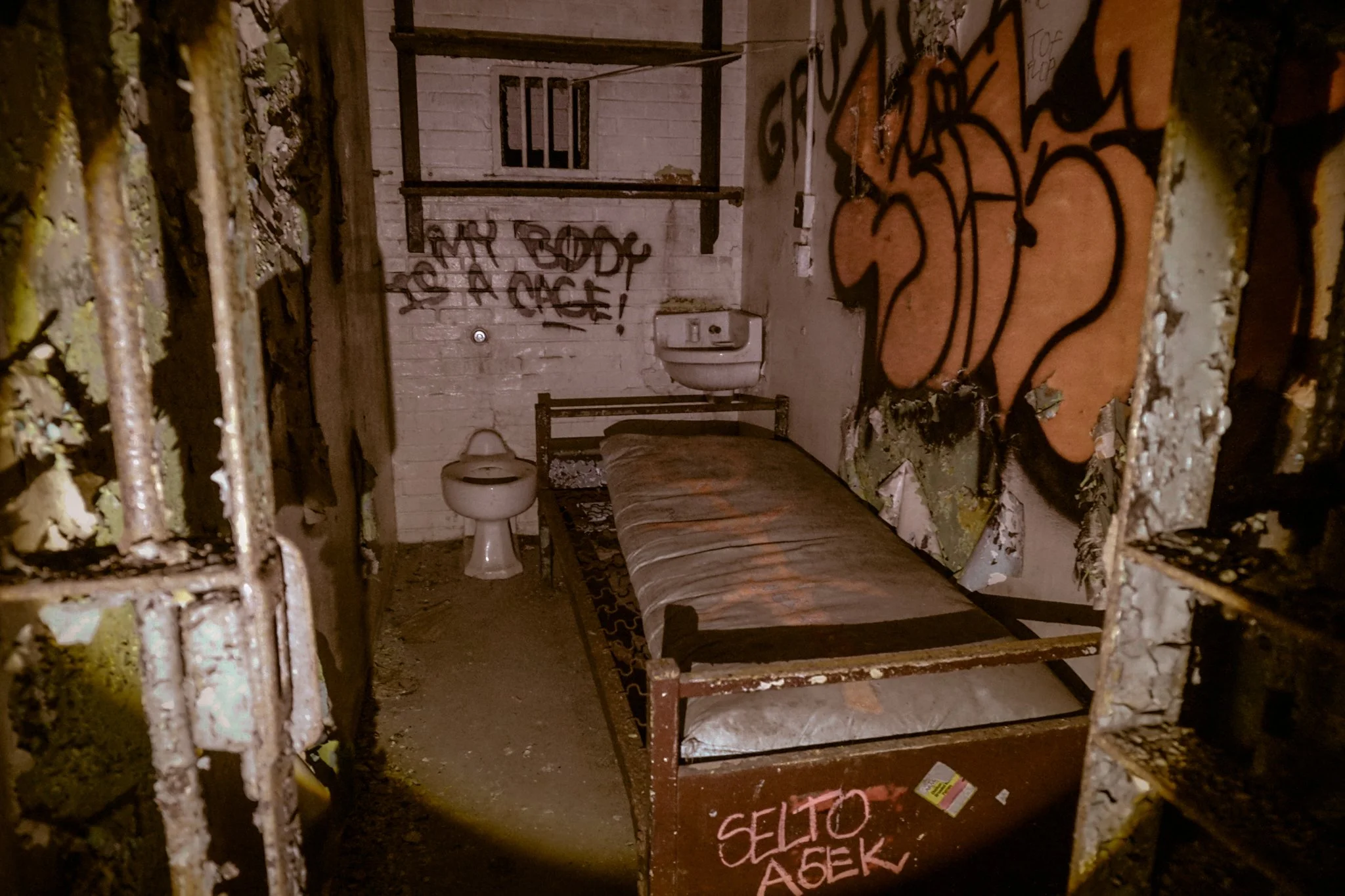 Abandoned jail cell with rusted metal bed frame, dirty mattress, graffiti on walls, and graffiti reading "SELTOT AEER" on the foot of the bed in a dimly lit room.