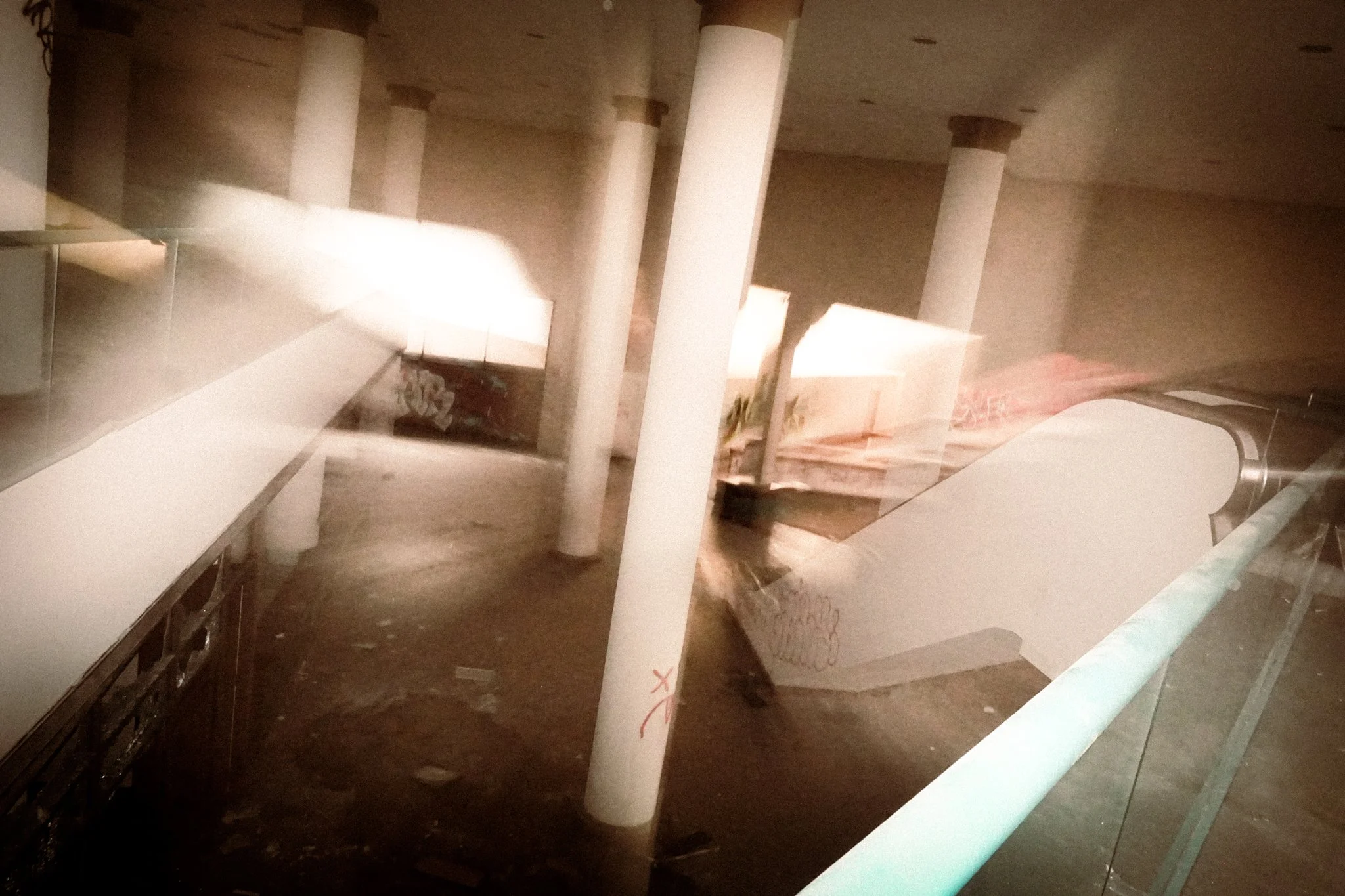 Interior of a building with white columns, a counter or desk, graffiti on a wall, and bright lights reflecting off a glass surface, with a blurred motion effect.