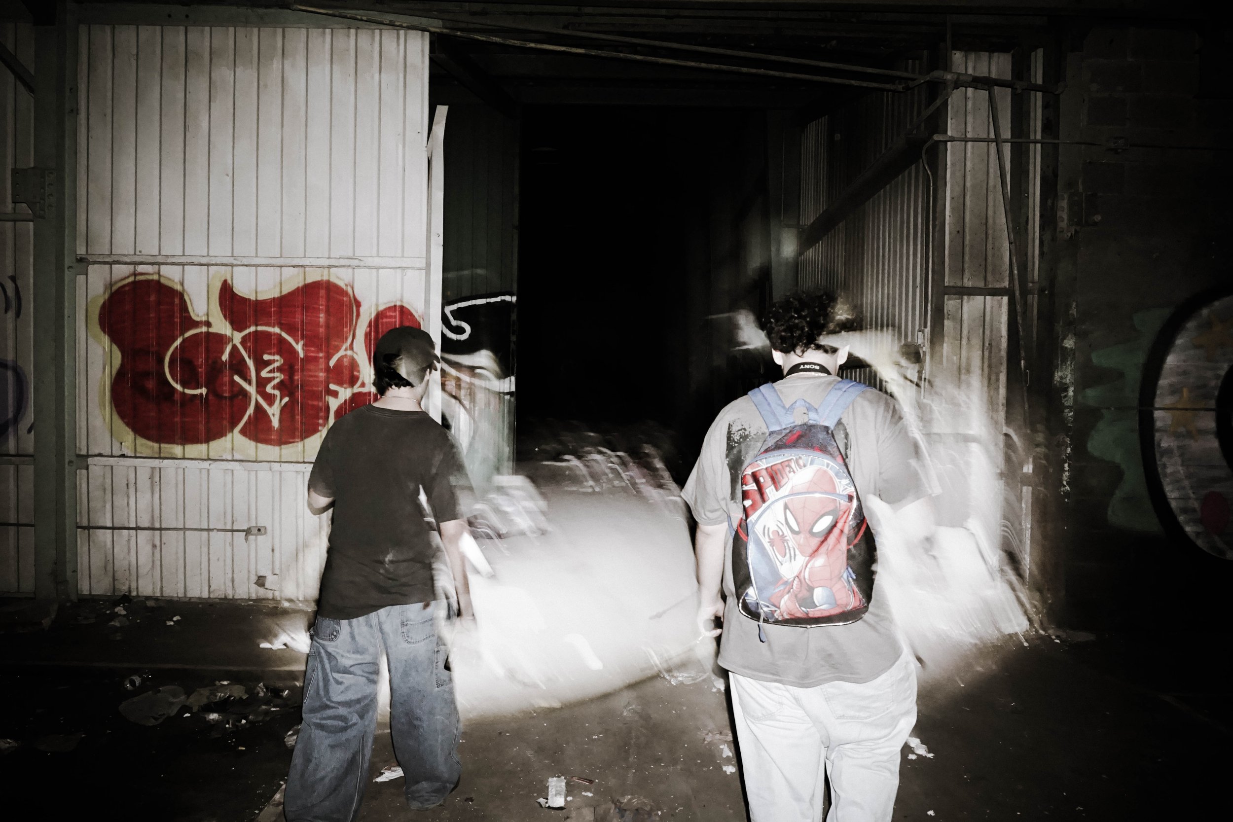 Two young people walking away from the camera in a dark, graffiti-covered alleyway. One is wearing a backpack with a Spider-Man design. The alley has metal walls, some graffiti, and debris on the ground.