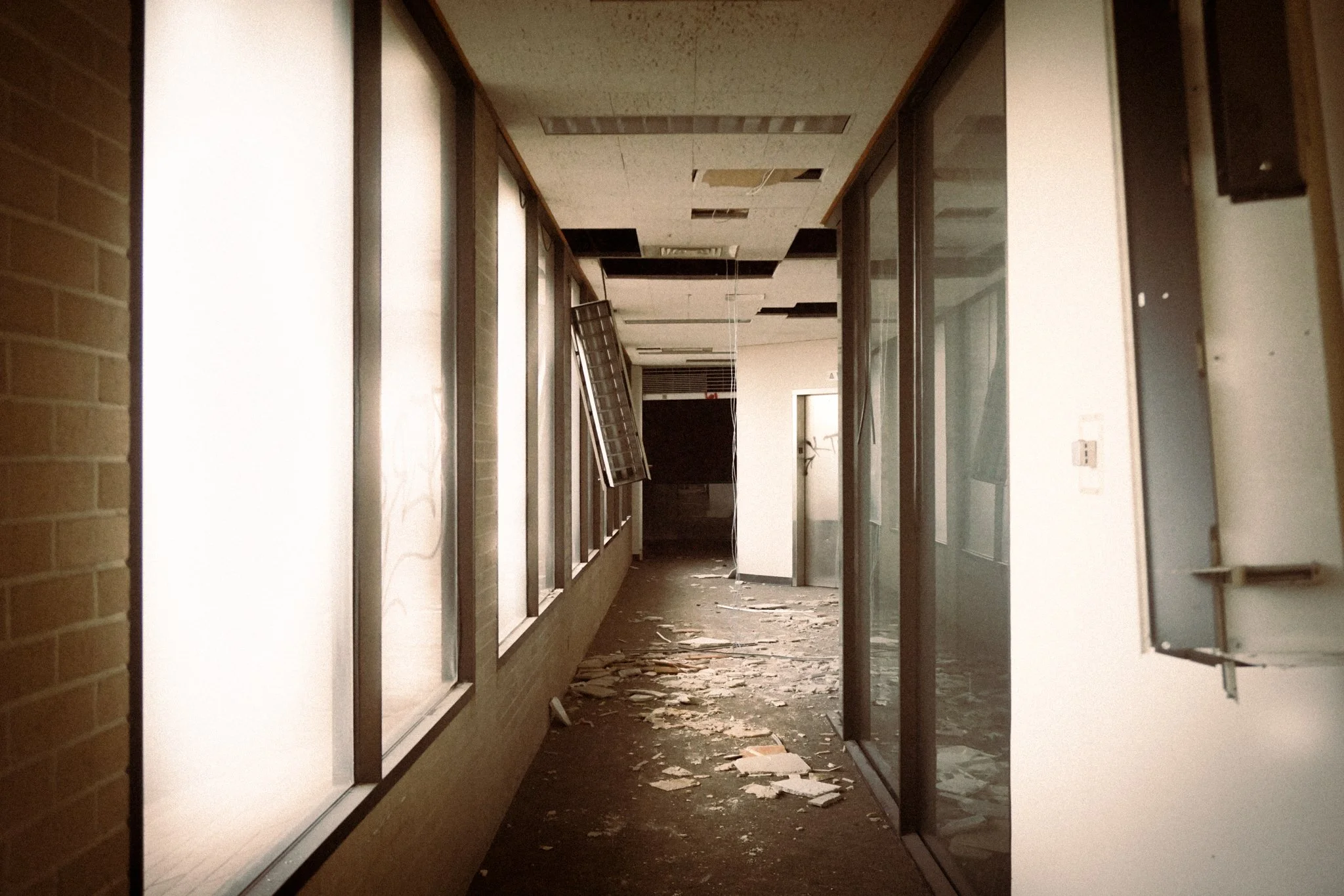Abandoned office hallway with broken ceiling tiles, scattered debris, and shattered windows.