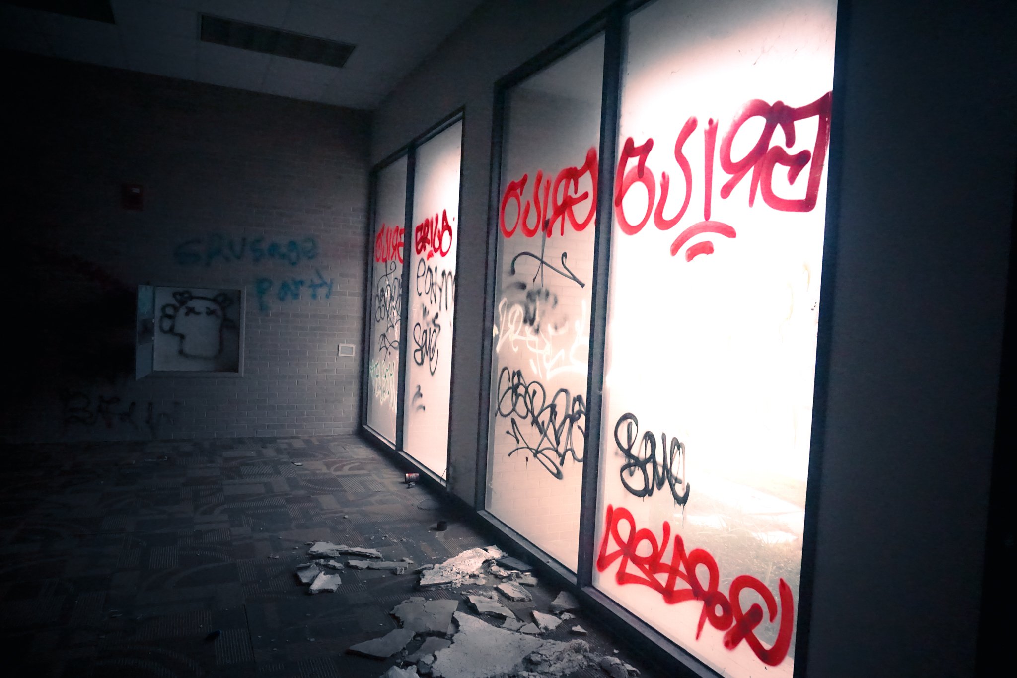 Graffiti-covered glass panels inside a dimly lit, abandoned building with debris on the floor.