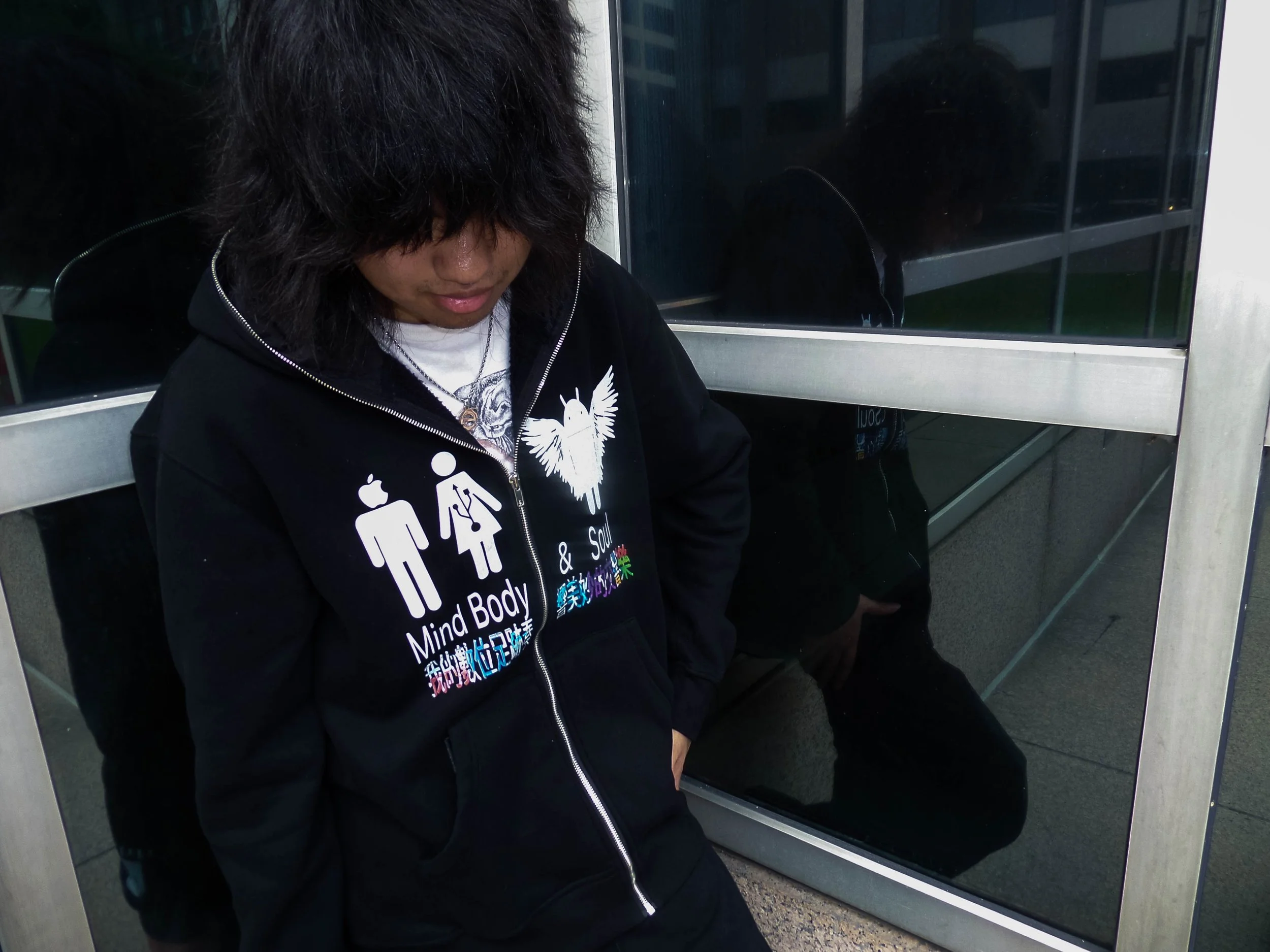 Person with curly black hair wearing a black jacket with graphic text, standing outside near glass doors.