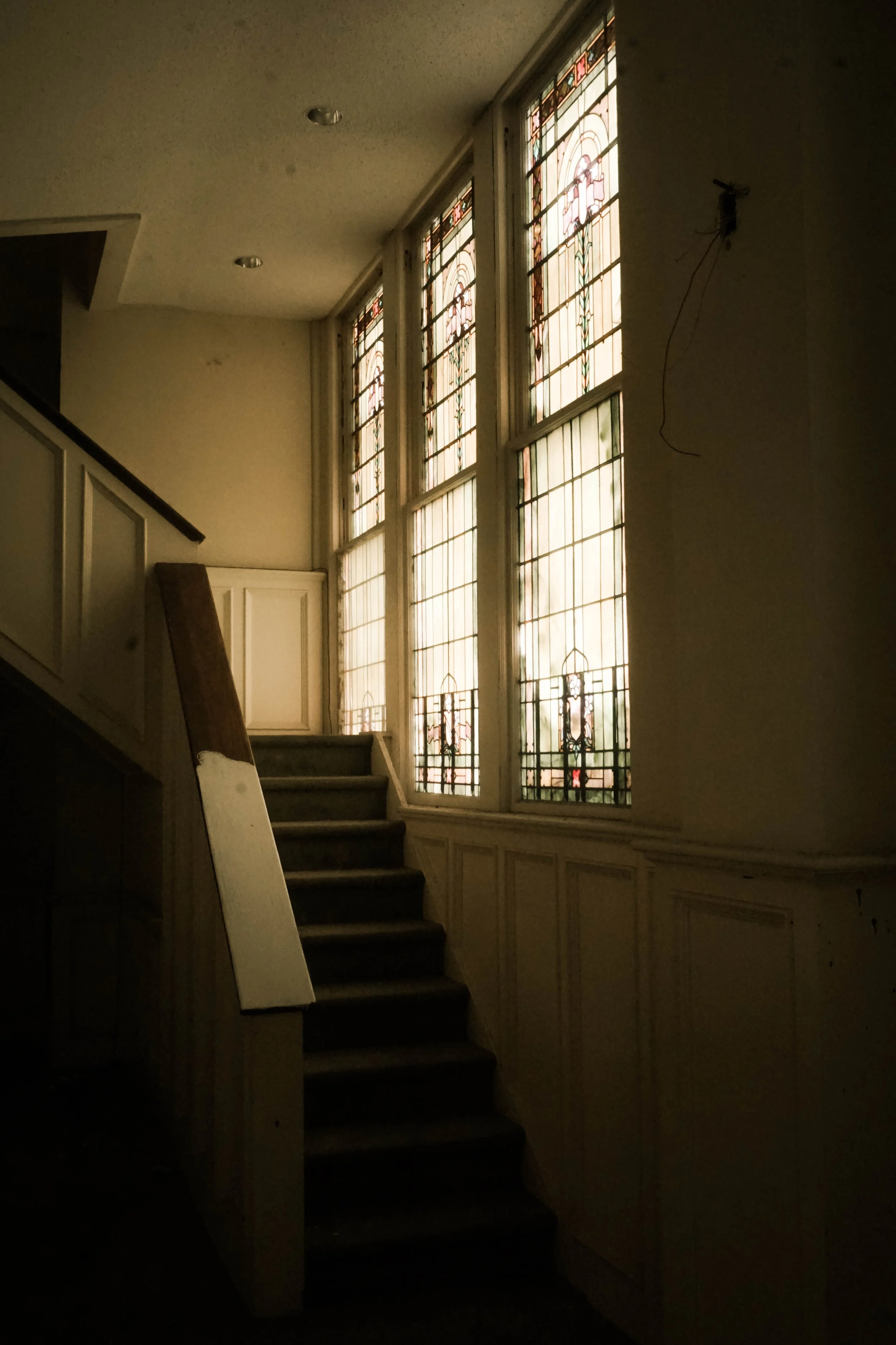 Dark staircase inside a building with stained glass windows on the right side, some wall paneling, and an unfinished electrical outlet on the wall.