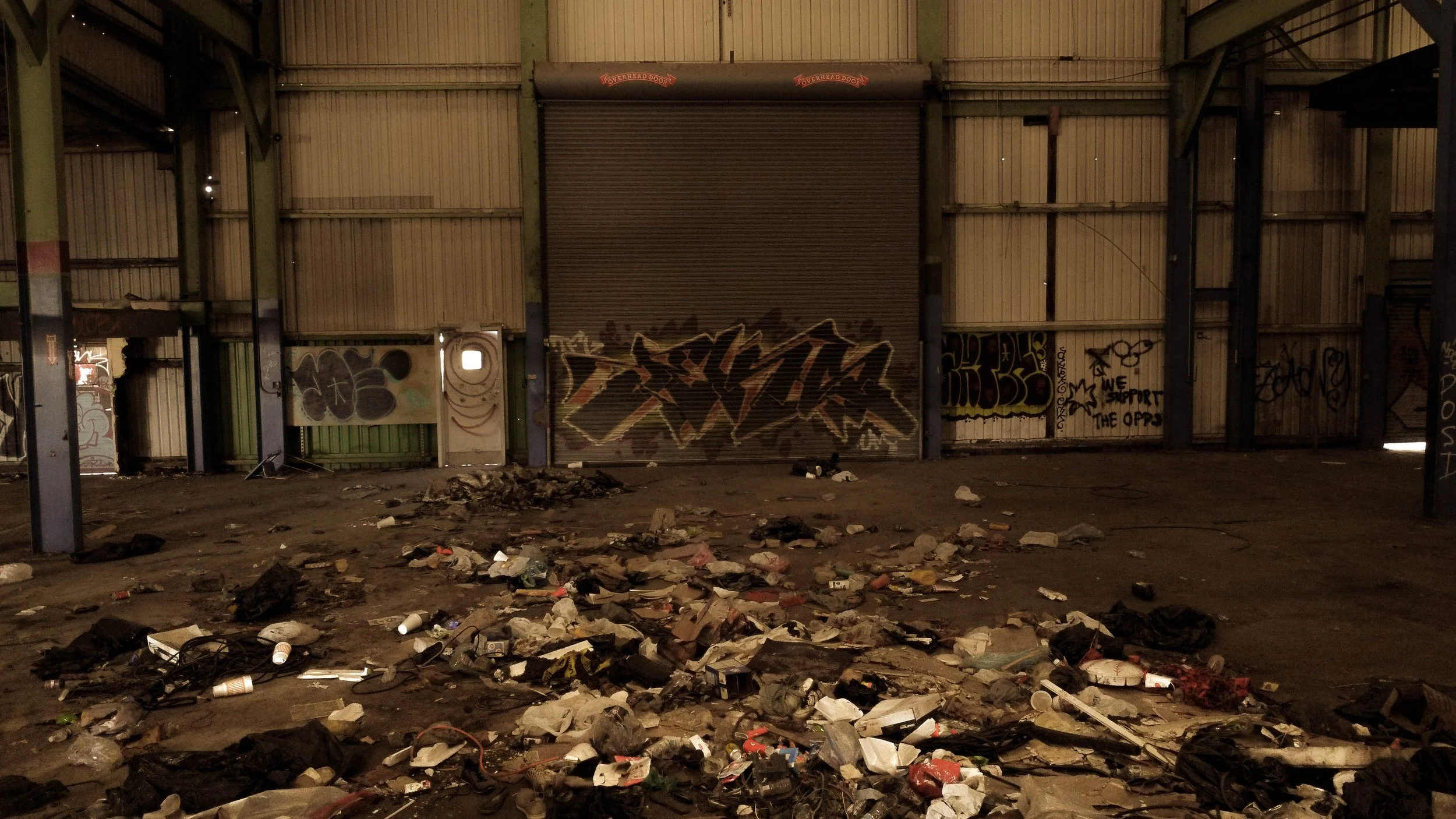 An abandoned, graffiti-covered warehouse with trash and debris scattered on the ground.