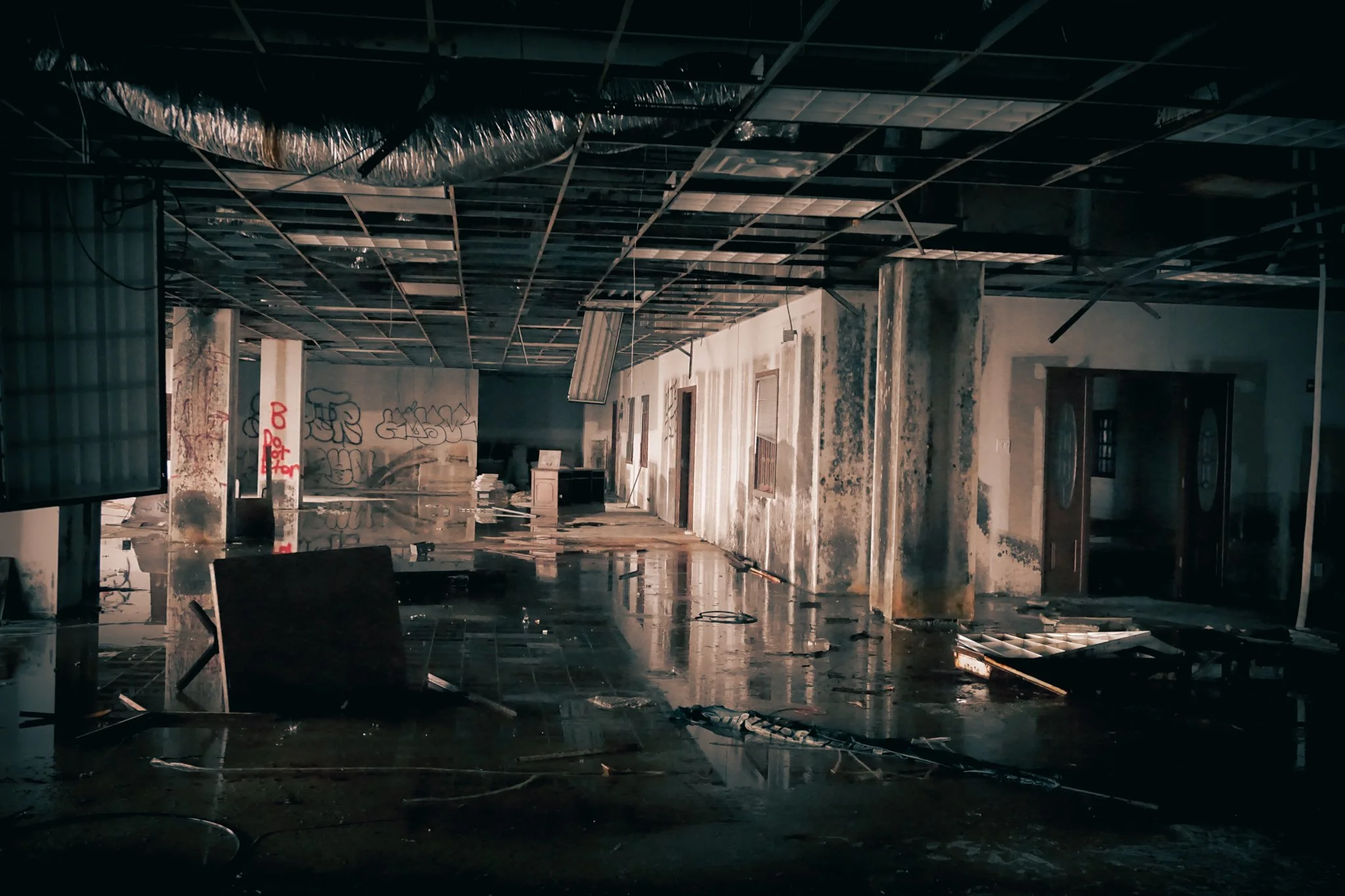 Interior of a damaged building with debris, peeling walls, and a partially collapsed ceiling, with graffiti on a column.