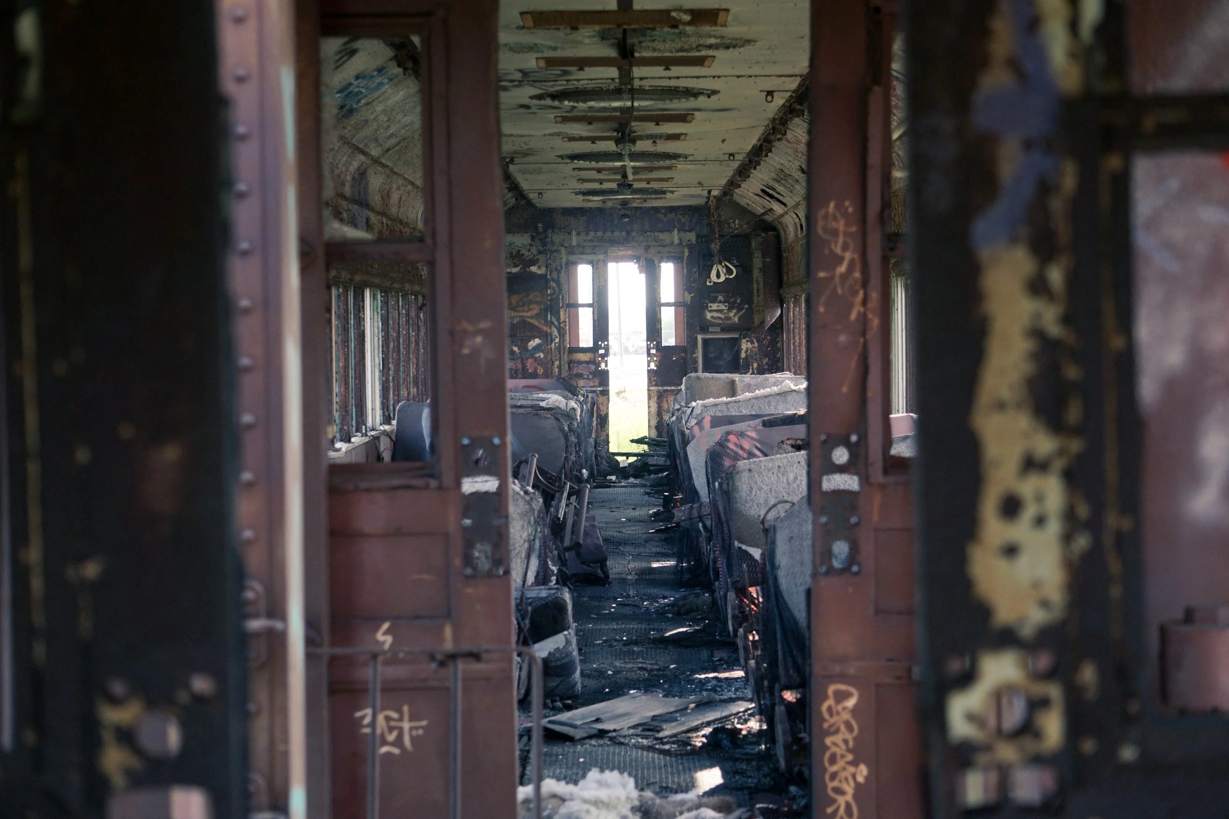 Inside a burned-out bus with charred seats and debris, viewed through an open door, showing the damaged interior and windows at the far end.