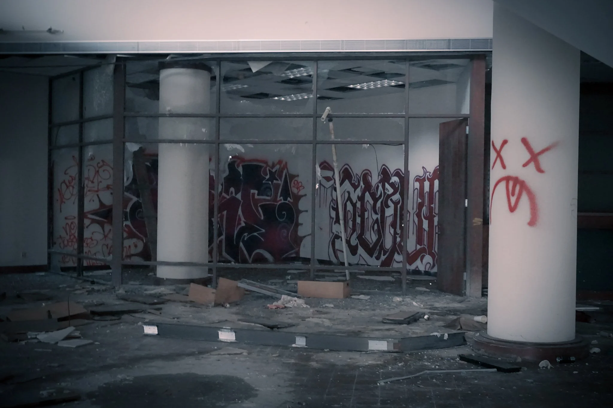 An abandoned, dilapidated room with broken glass, graffitied walls, and scattered debris on the floor, surrounded by metal framing.