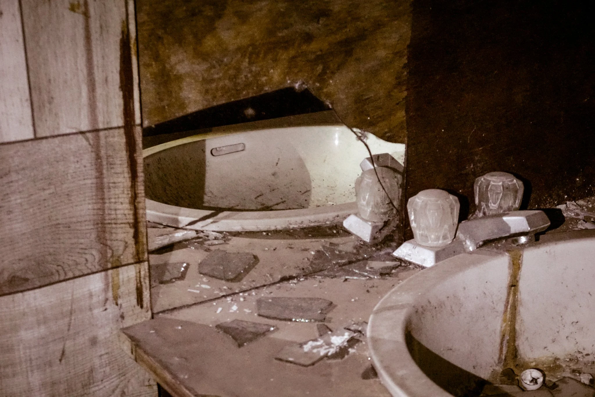 Broken and dirty bathroom with abandoned washing machine and sink, debris, and shattered glass.