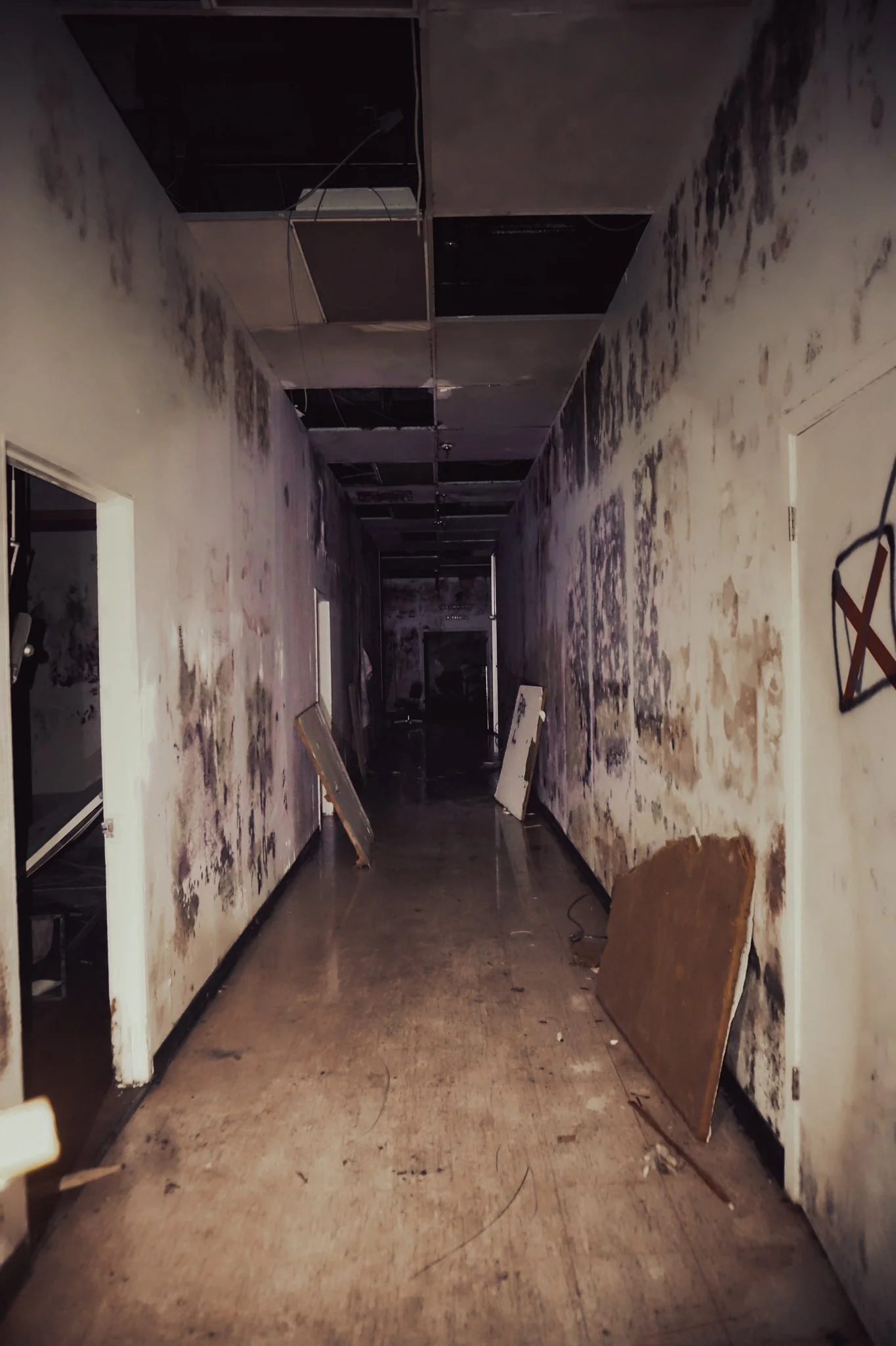 A long abandoned hallway with water-stained walls, a damaged ceiling with missing tiles, and debris including a fallen board and broken frames on the floor.