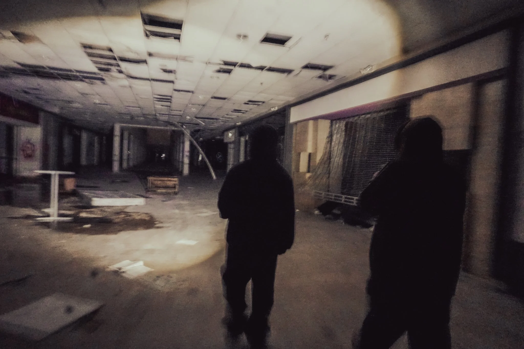Two silhouetted figures walk through an abandoned, damaged indoor space with a broken ceiling and debris scattered on the floor.