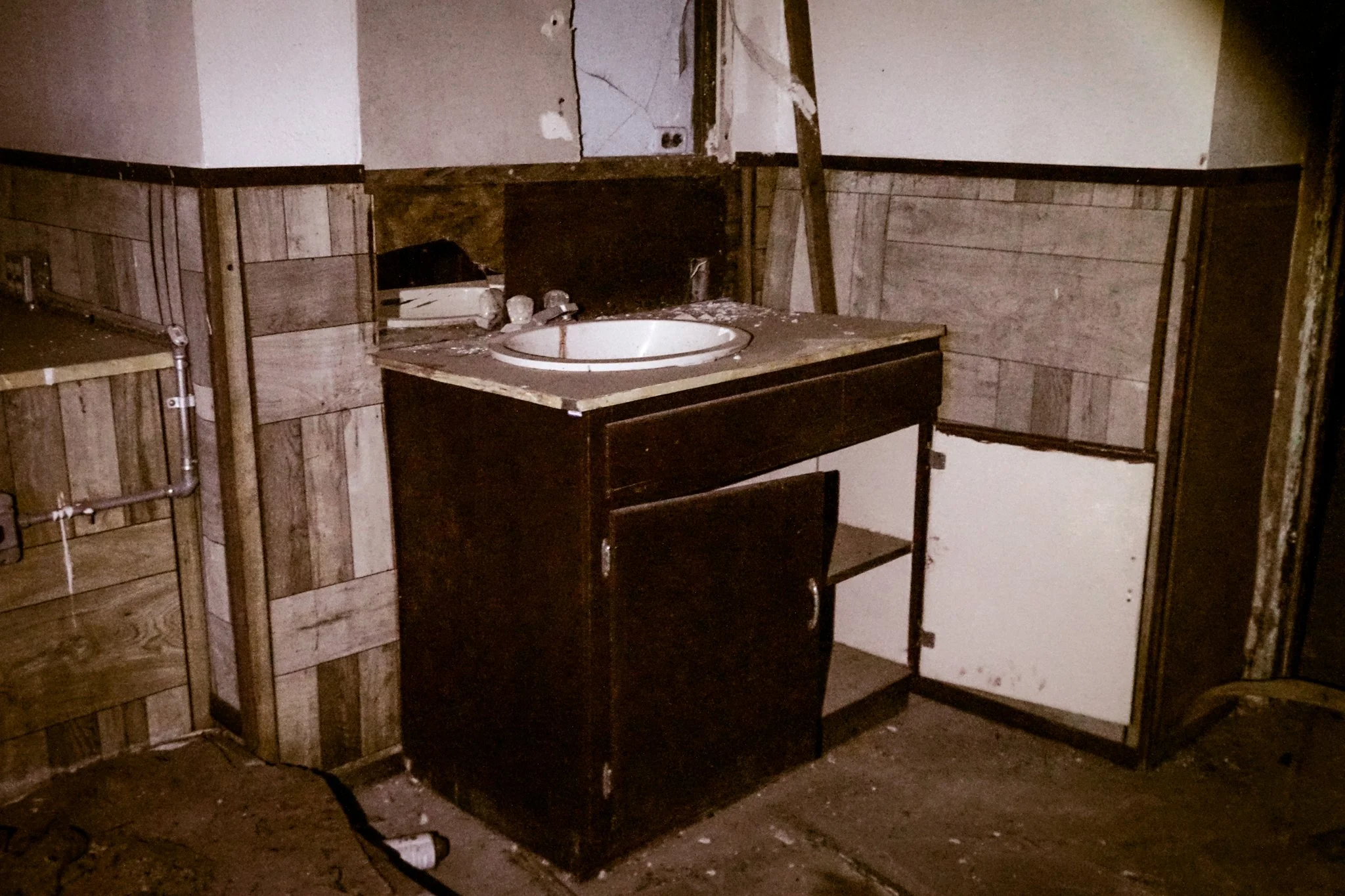 Dirty bathroom with a dark wooden cabinet, a white sink, and unfinished wood and drywall walls in a state of disrepair.