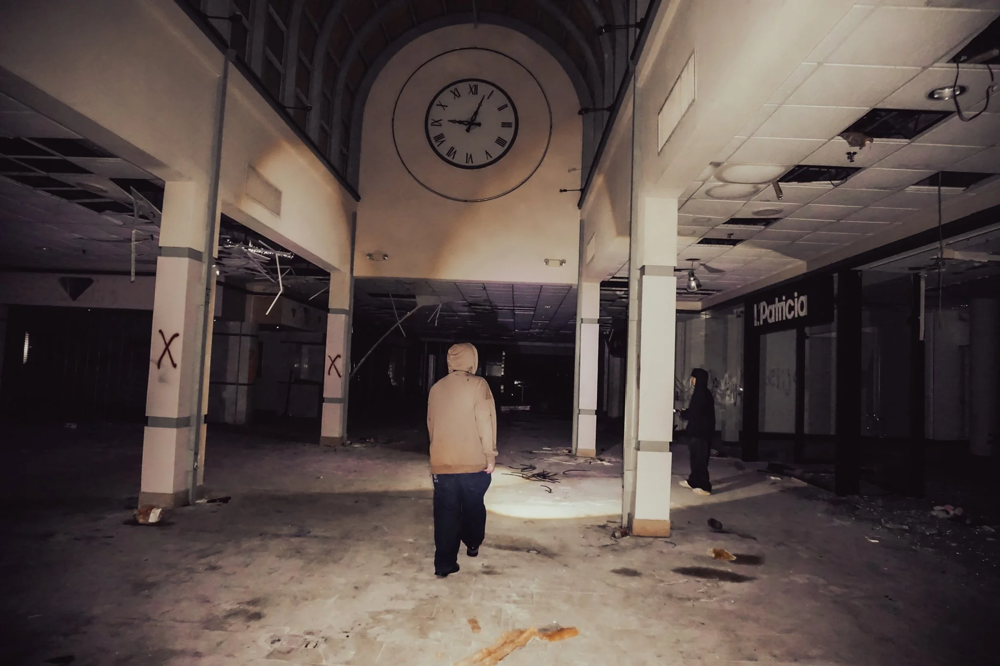 An abandoned shopping mall with a large clock on the wall showing 7:58. The mall appears damaged, with missing ceiling tiles and debris on the floor. Two people are walking through the space, and closed stores are visible, including one named 'L. Pat