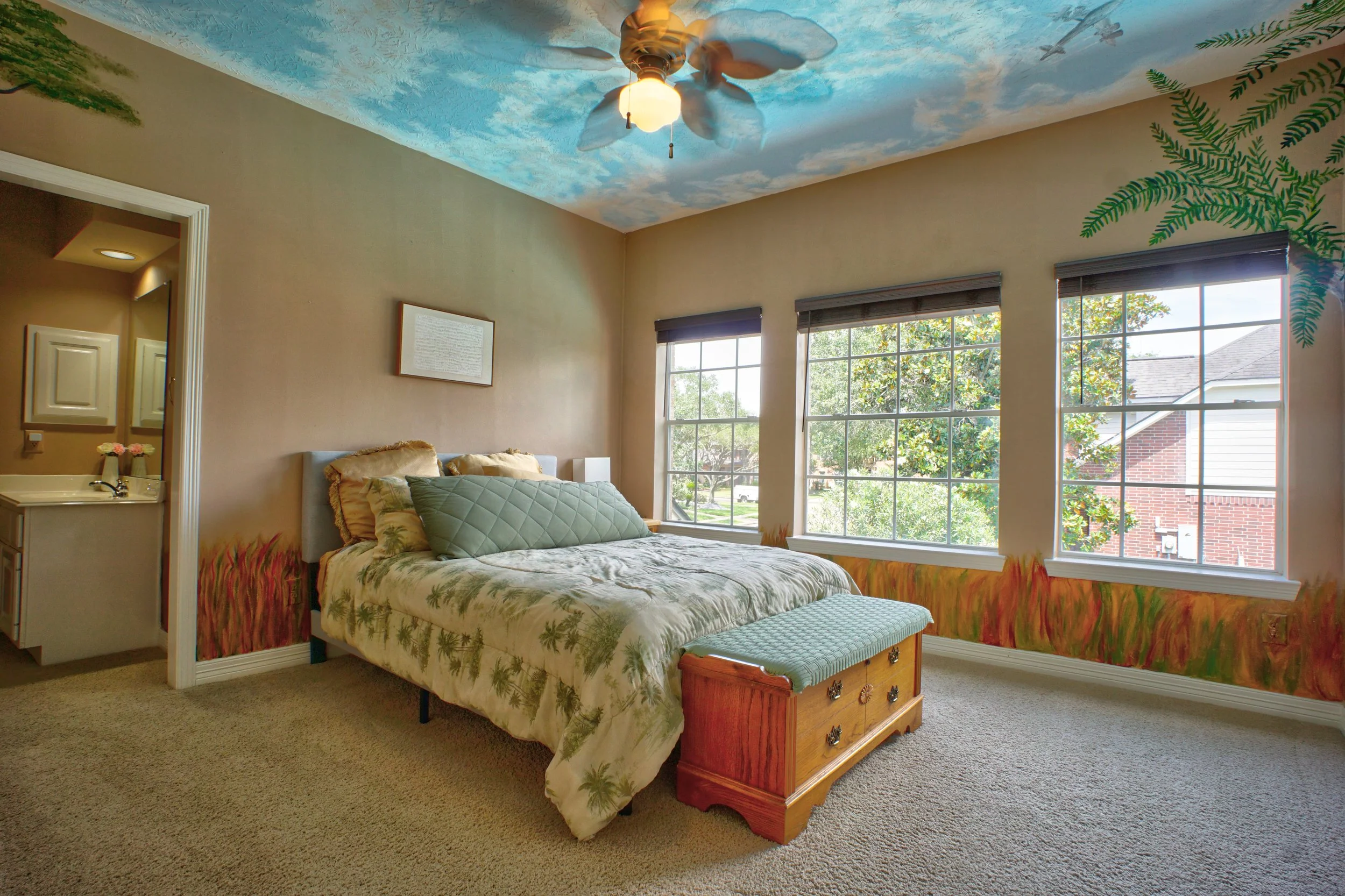 A bedroom with three large windows showing green trees outside, a bed with palm tree patterned bedding, a wooden chest at the foot of the bed, and a ceiling painted like a sky with clouds and a ceiling fan.