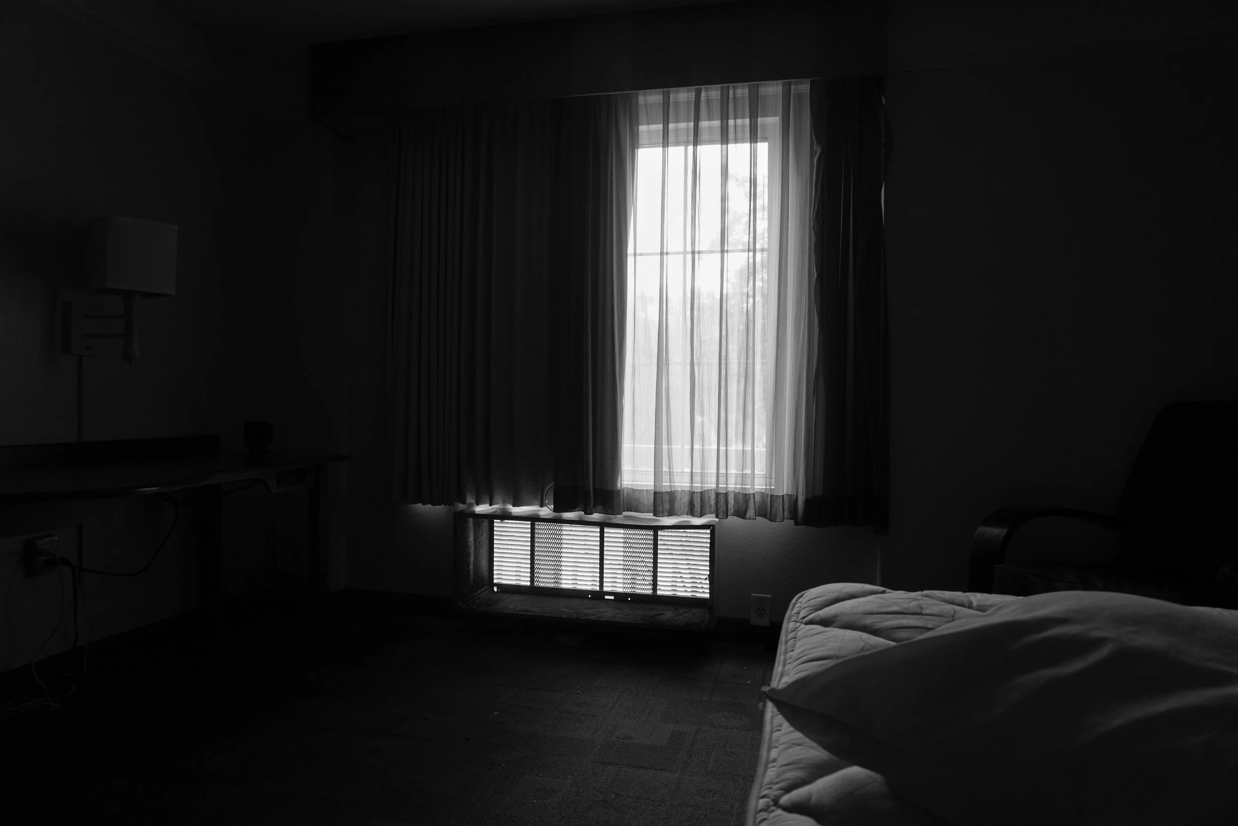 A dark hotel room with a window covered by semi-transparent curtains, sunlight filtering through, with a bed partly visible in the foreground, and a desk on the left side.