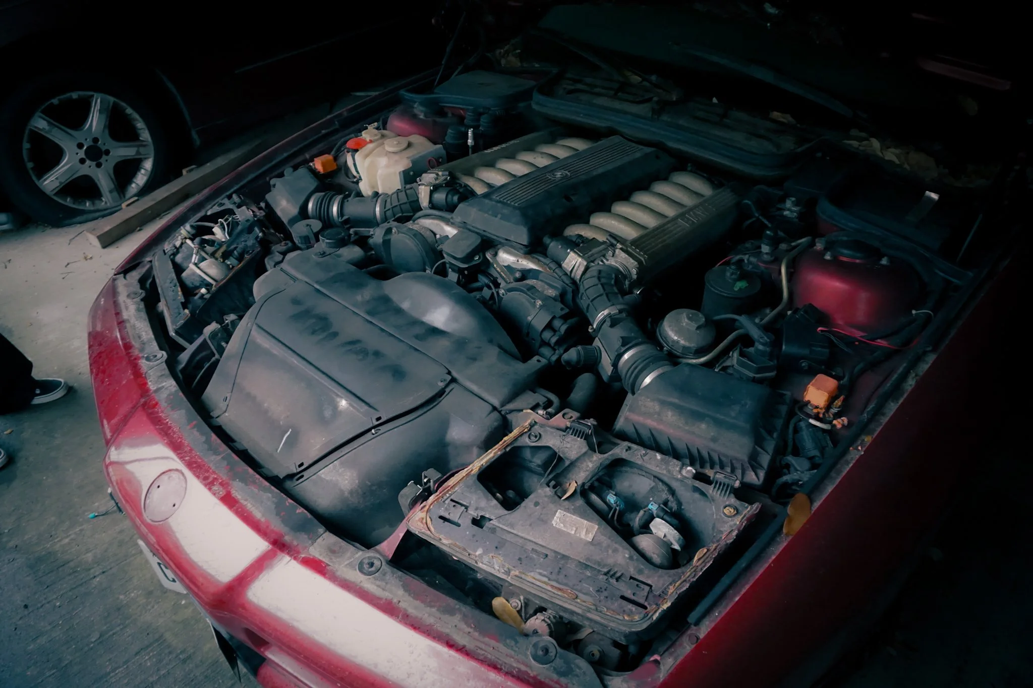 The open hood of a red car reveals the engine, with various mechanical parts visible, in a garage with a car wheel on a stand nearby.