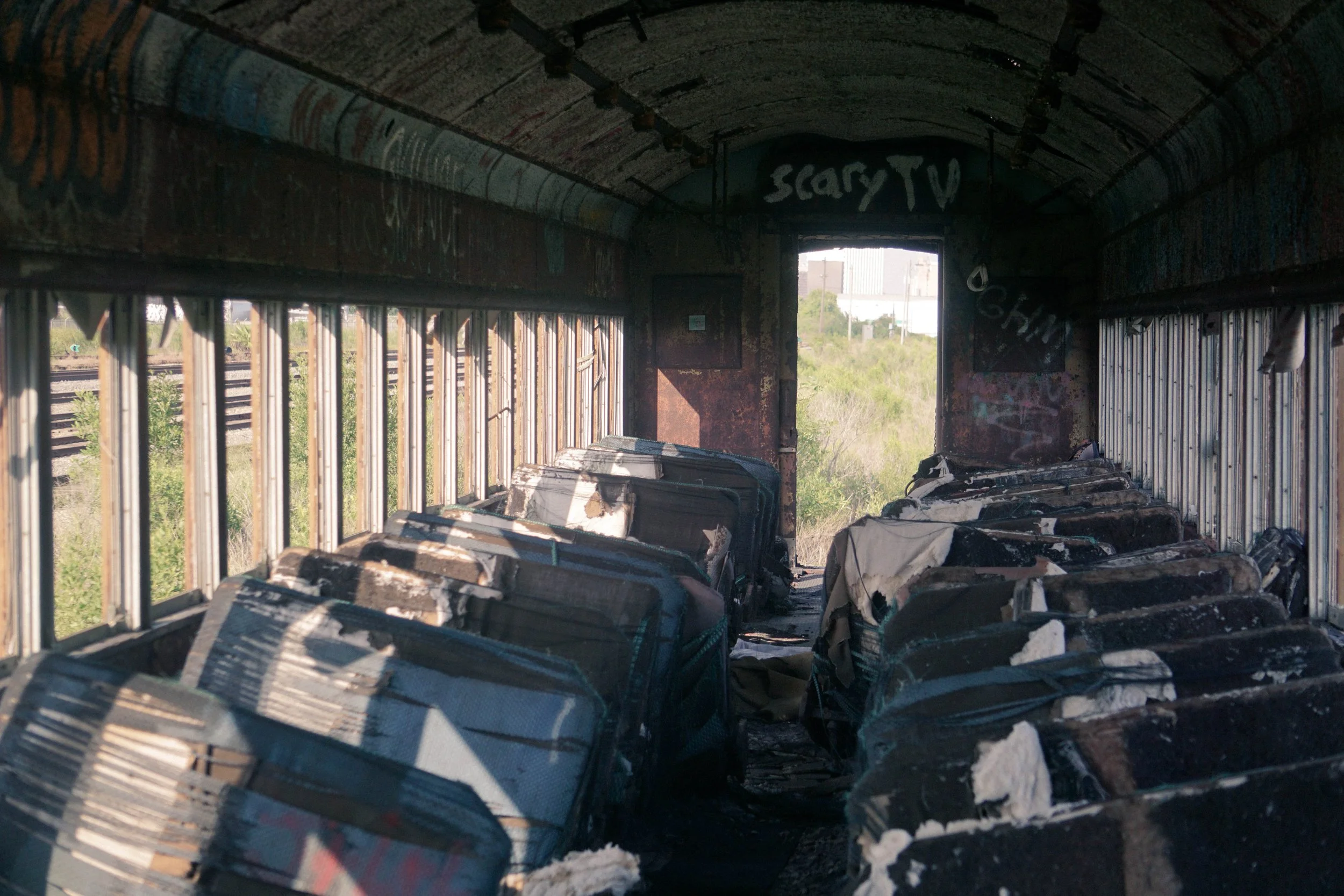 Inside an abandoned bus, with burnt seats and graffiti on the walls and ceiling, overlooking a landscape with grass and buildings outside.