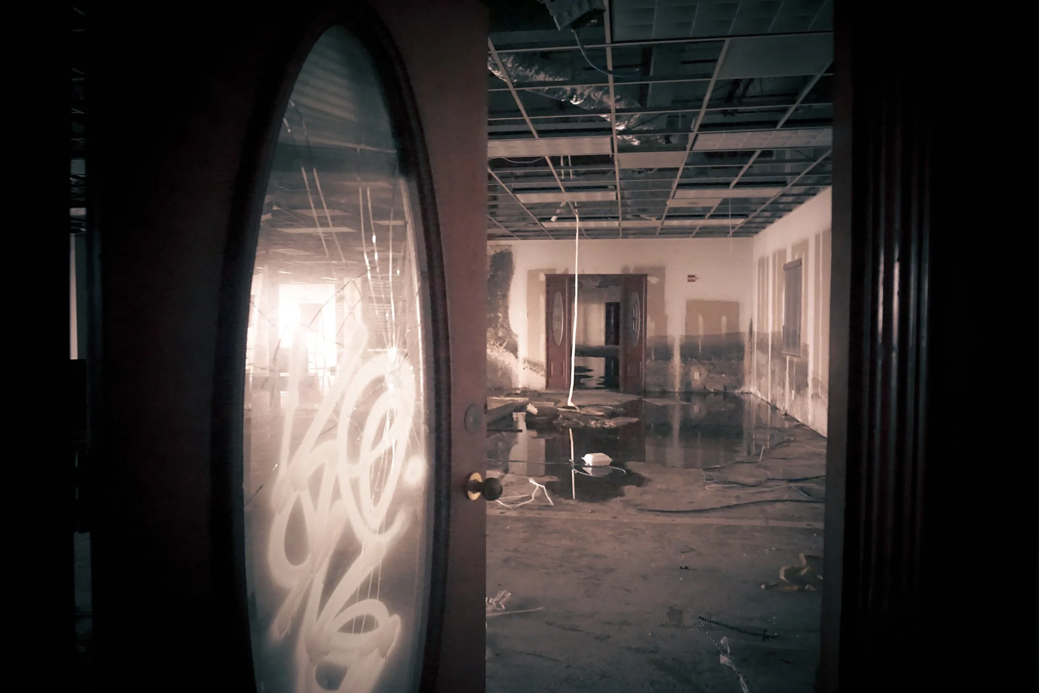An interior space of a building under construction or renovation, showing a damaged or burned wall, a flooded floor, hanging wires, and an unfinished ceiling with exposed framework. The image is taken from outside through a door with a decorative gla