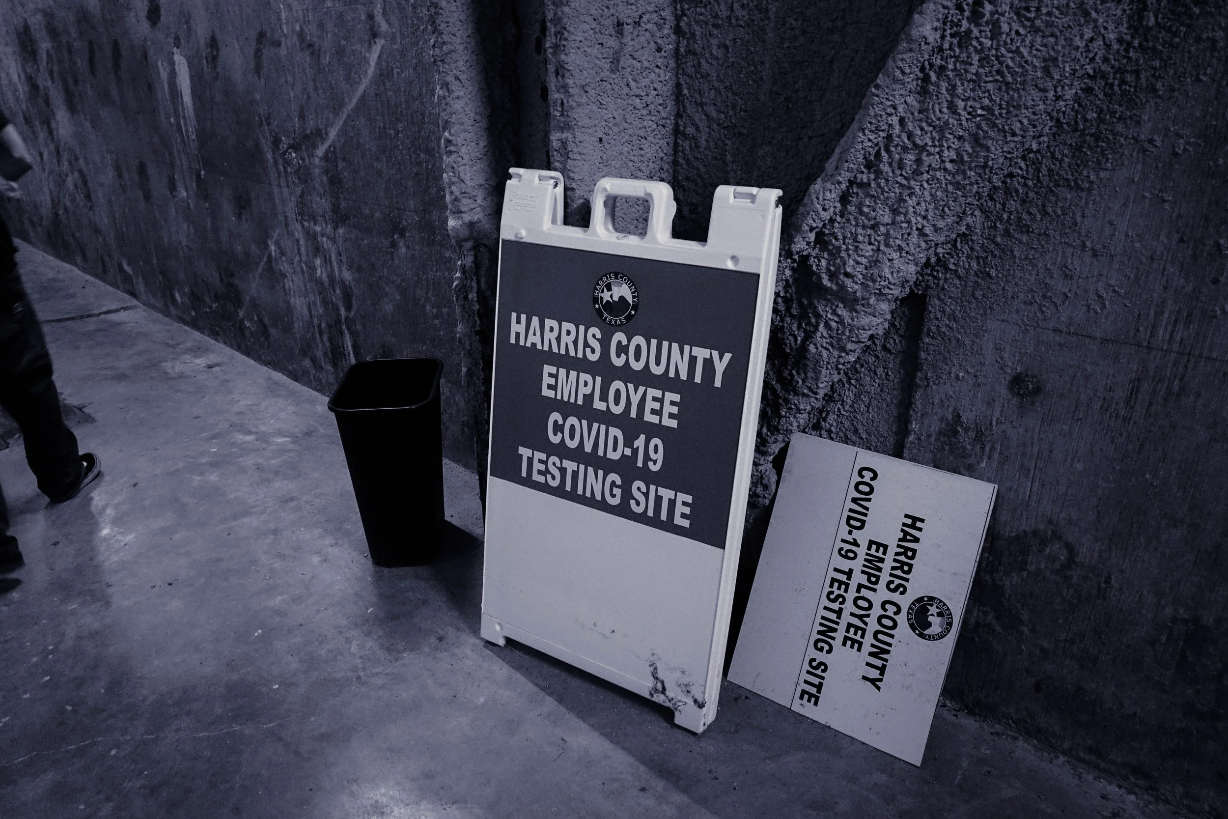 Sign for Harris County employee COVID-19 testing site leaning against a concrete wall, with a trash can nearby.