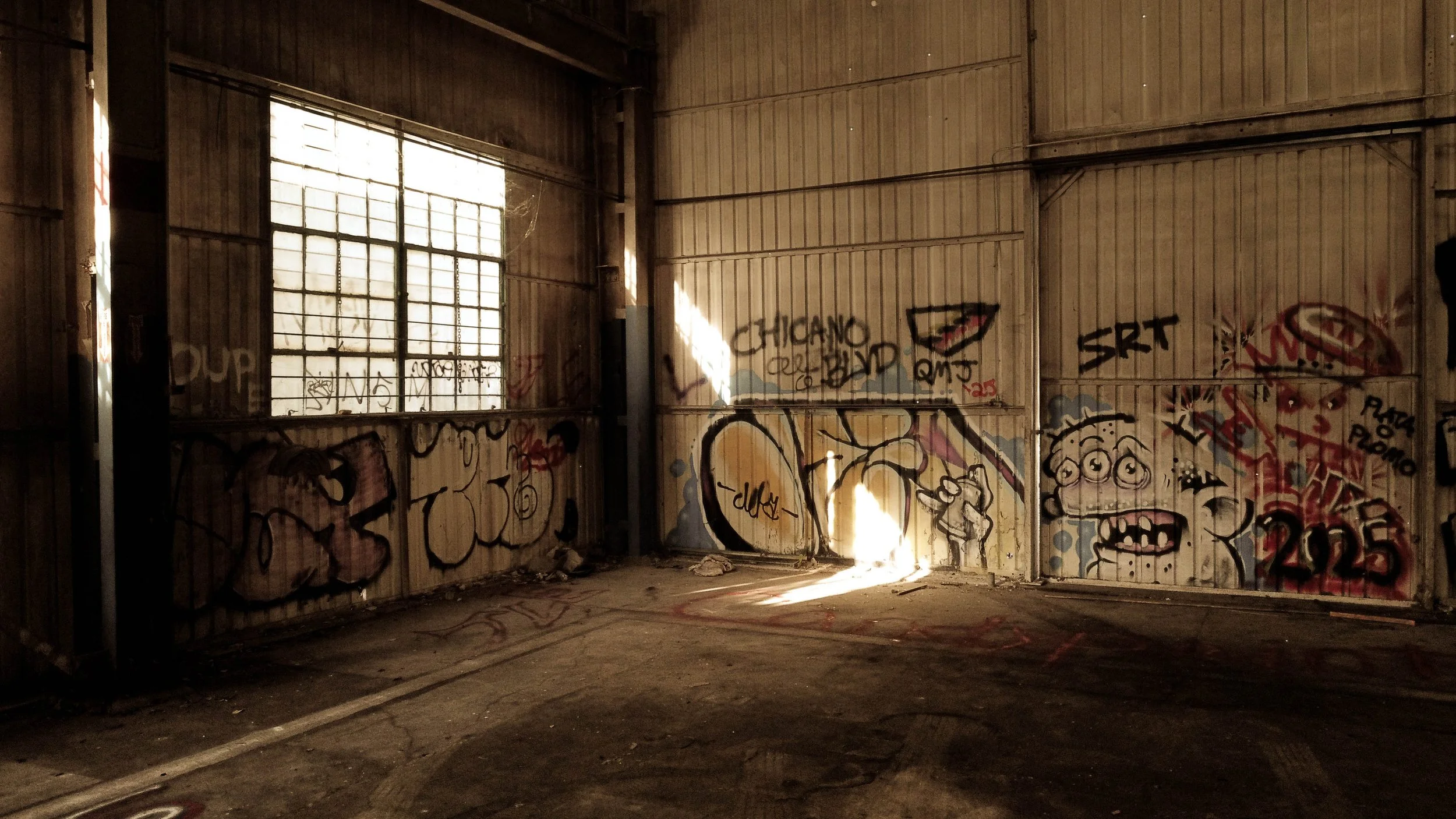 An abandoned, graffitied warehouse with a large window allowing sunlight to stream in, illuminating the dirty, cracked floor.
