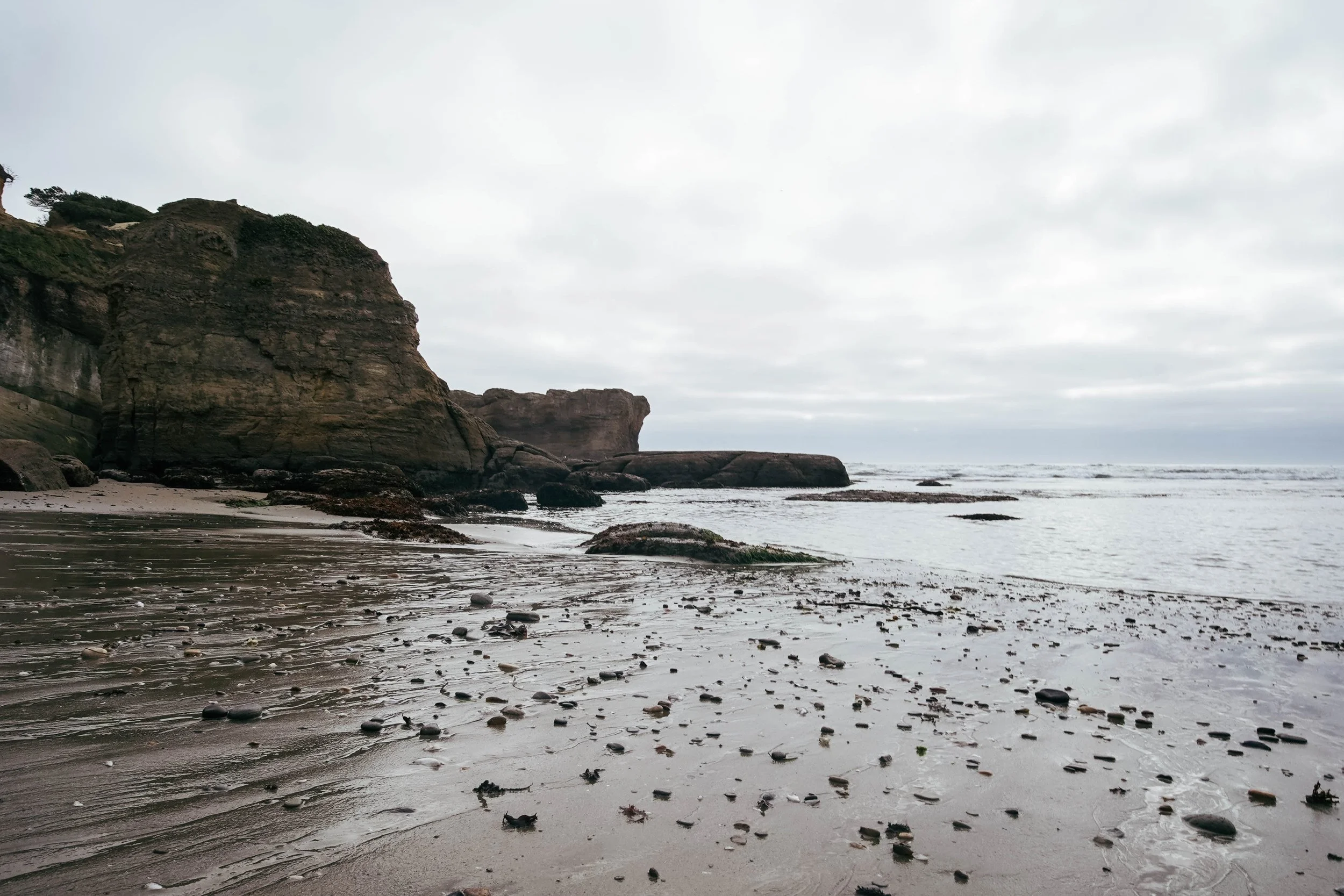 Overcast beach with rocky cliffs, wet sand, scattered stones, and gentle waves.