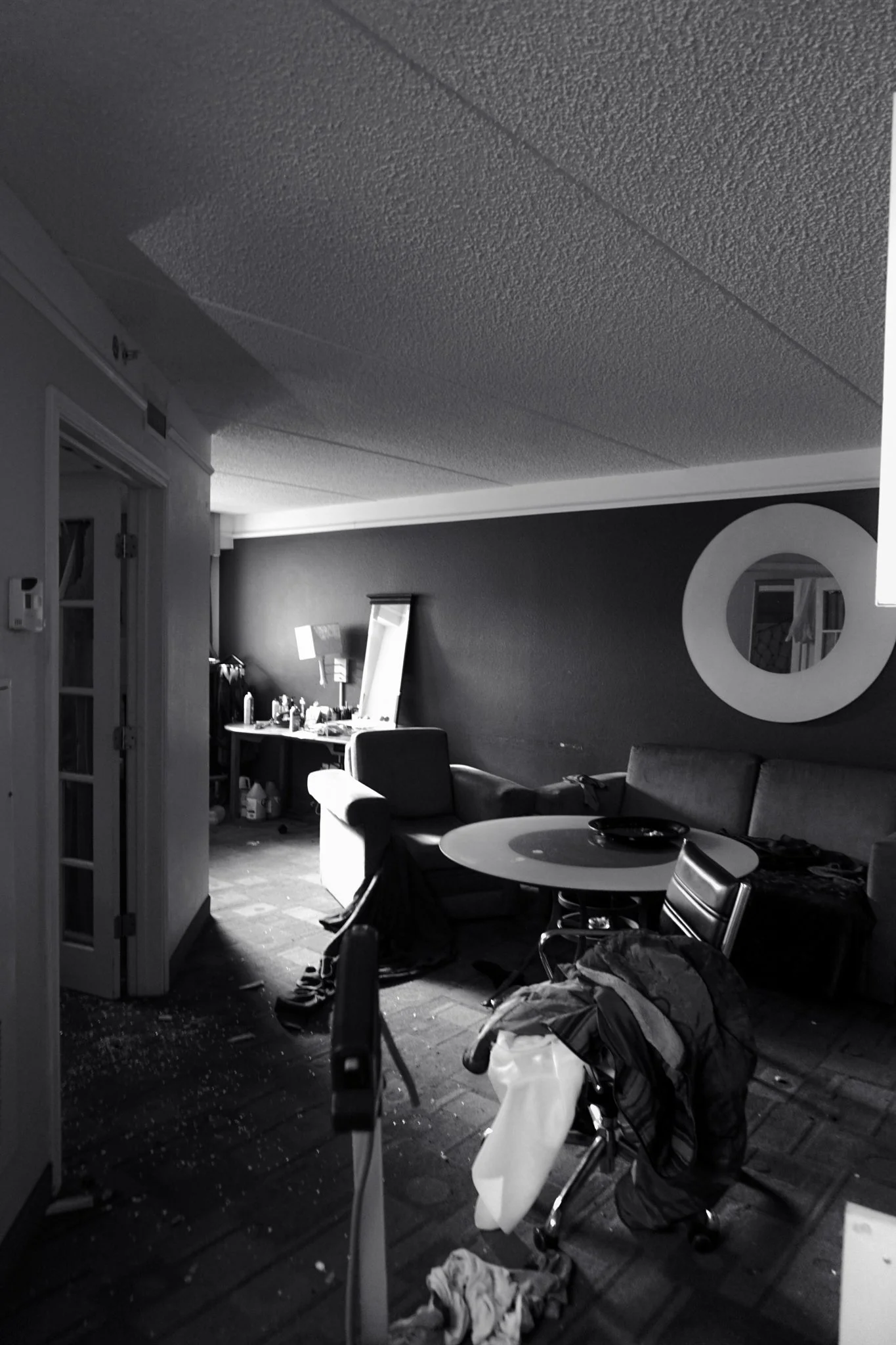 A messy living room with scattered clothes, a chair with clothing draped over it, a table with items on top, a sofa, and a desk cluttered with bottles and other objects, illuminated by a window.