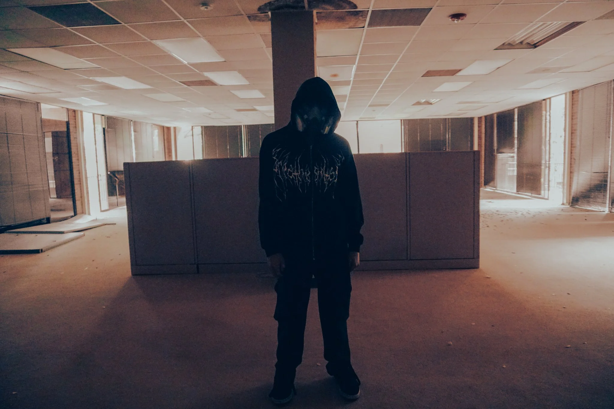 Person in dark clothing with hoodie standing in an empty, abandoned office space with broken blinds and dusty floors, backlit by sunlight through windows.