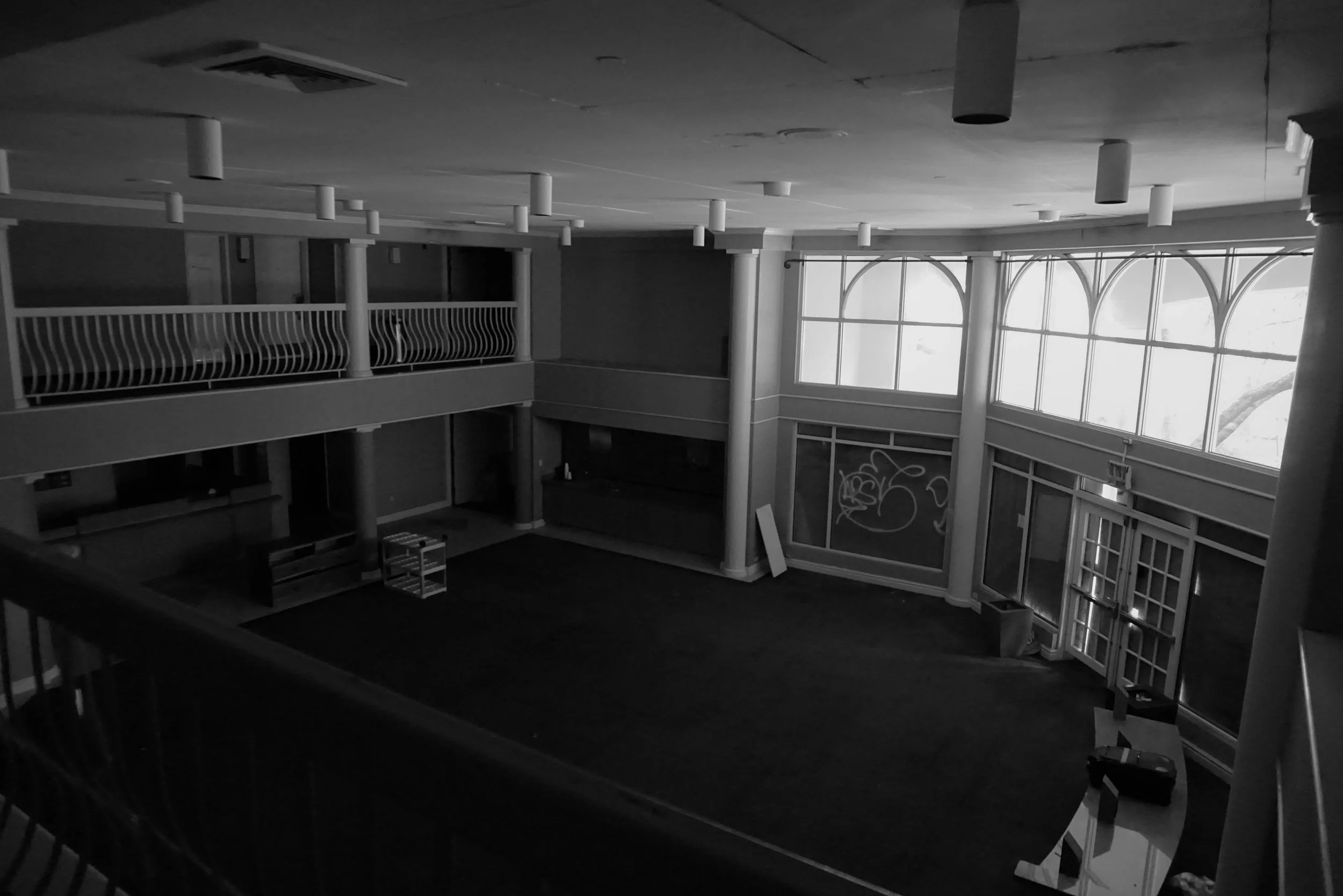 Empty interior space with large windows, some graffiti on the wall, and a second-floor balcony with railing.